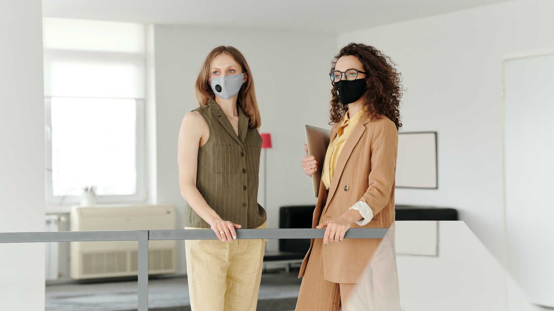 Two female leaders wearing face masks and looking over railing in modern office space