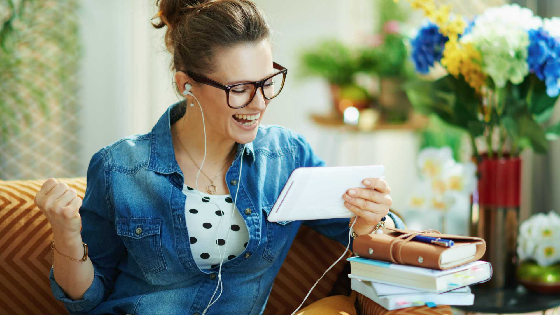 happy stylish female in jeans shirt with white headphones and tablet PC rejoicing while study online in the modern house in sunny day.