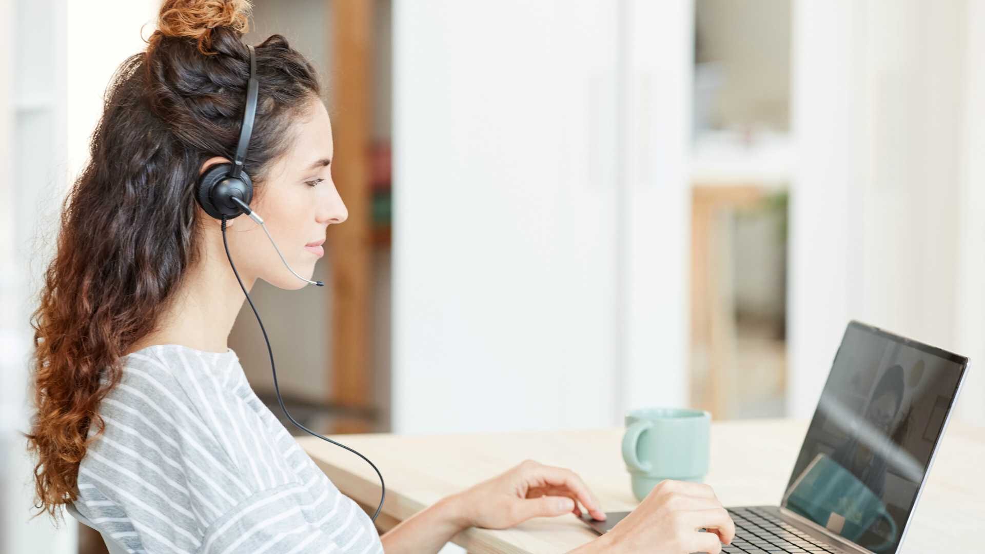 Modern young Caucasian woman wearing headset working on laptop at home, horizontal side view shot