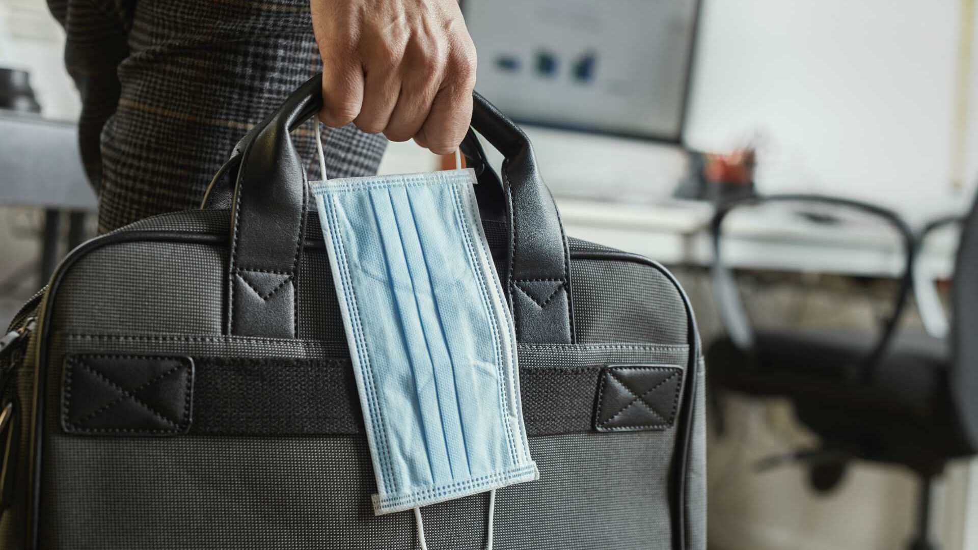 Closeup of a young man in an office holding a briefcase and a surgical mask in his hand