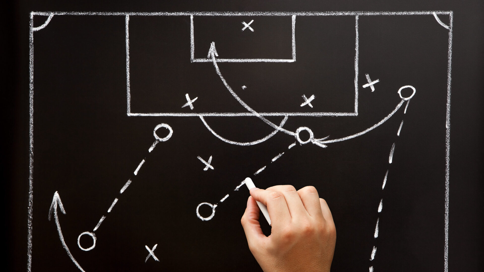 a chalkboard background with x's and o's arranged like a sport plan. A hand holds chalk in the bottom of the photo, arranging the shapes to improve performance.