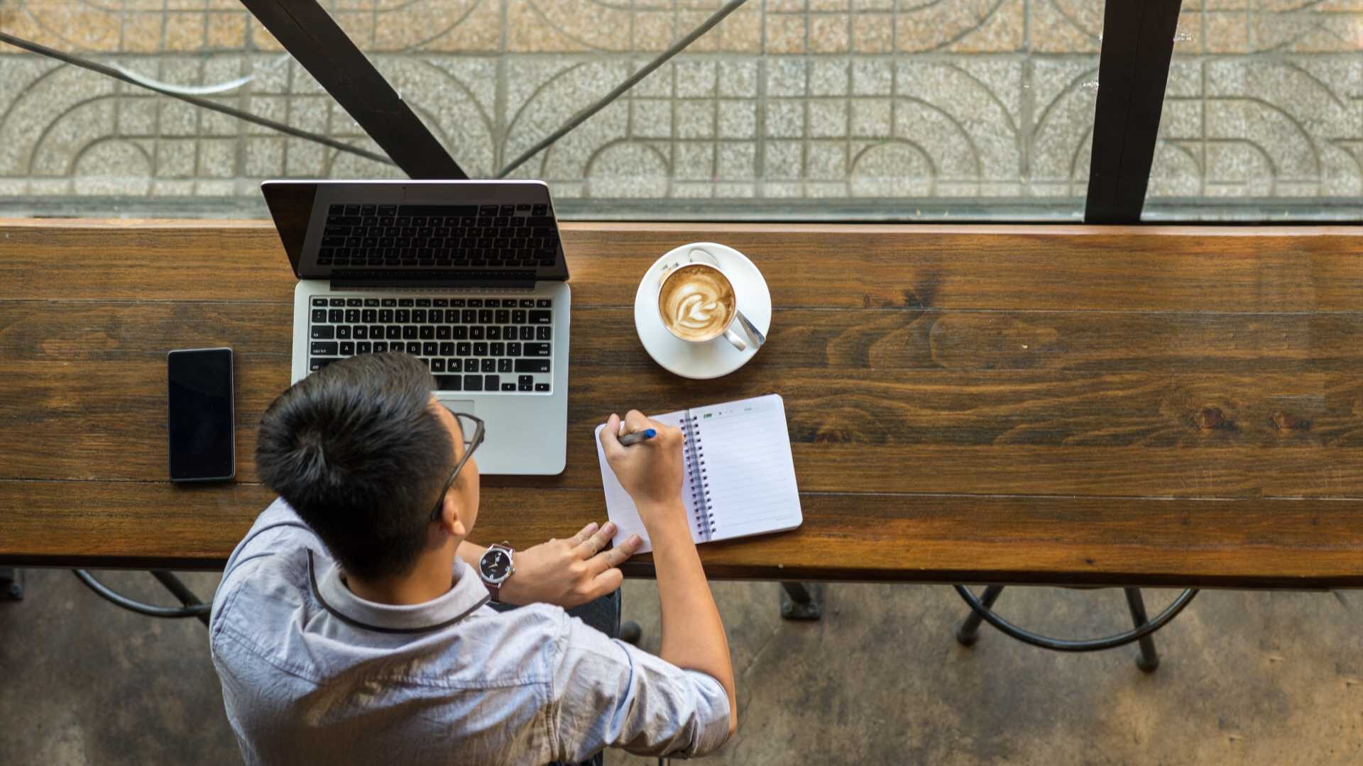 overhead shot of a young person working on their laptop at a coffee shop, they are writing notes in a notebook and enjoying a latte