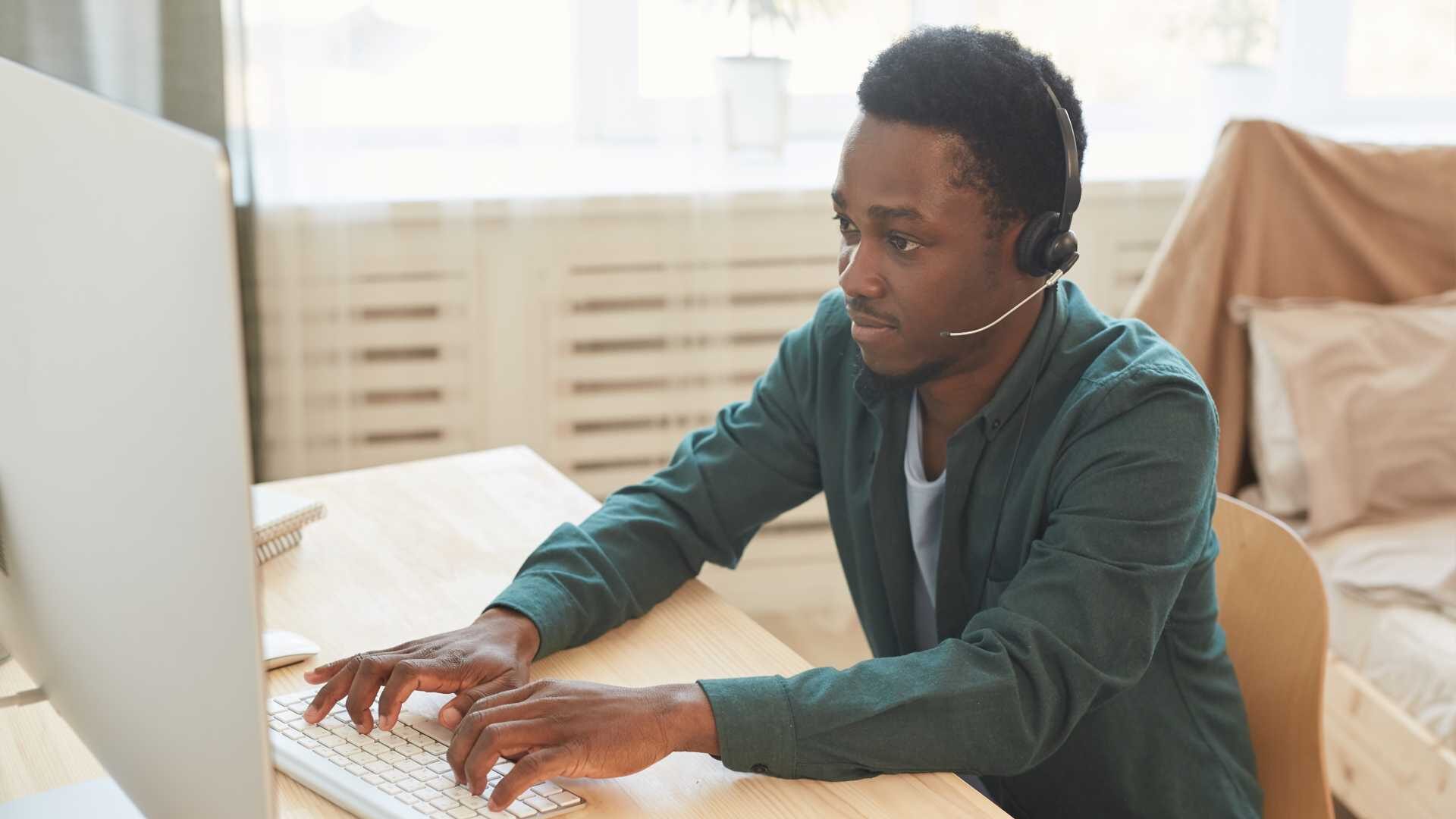 A young Black salesperson is typing on his computer with a headset on,
