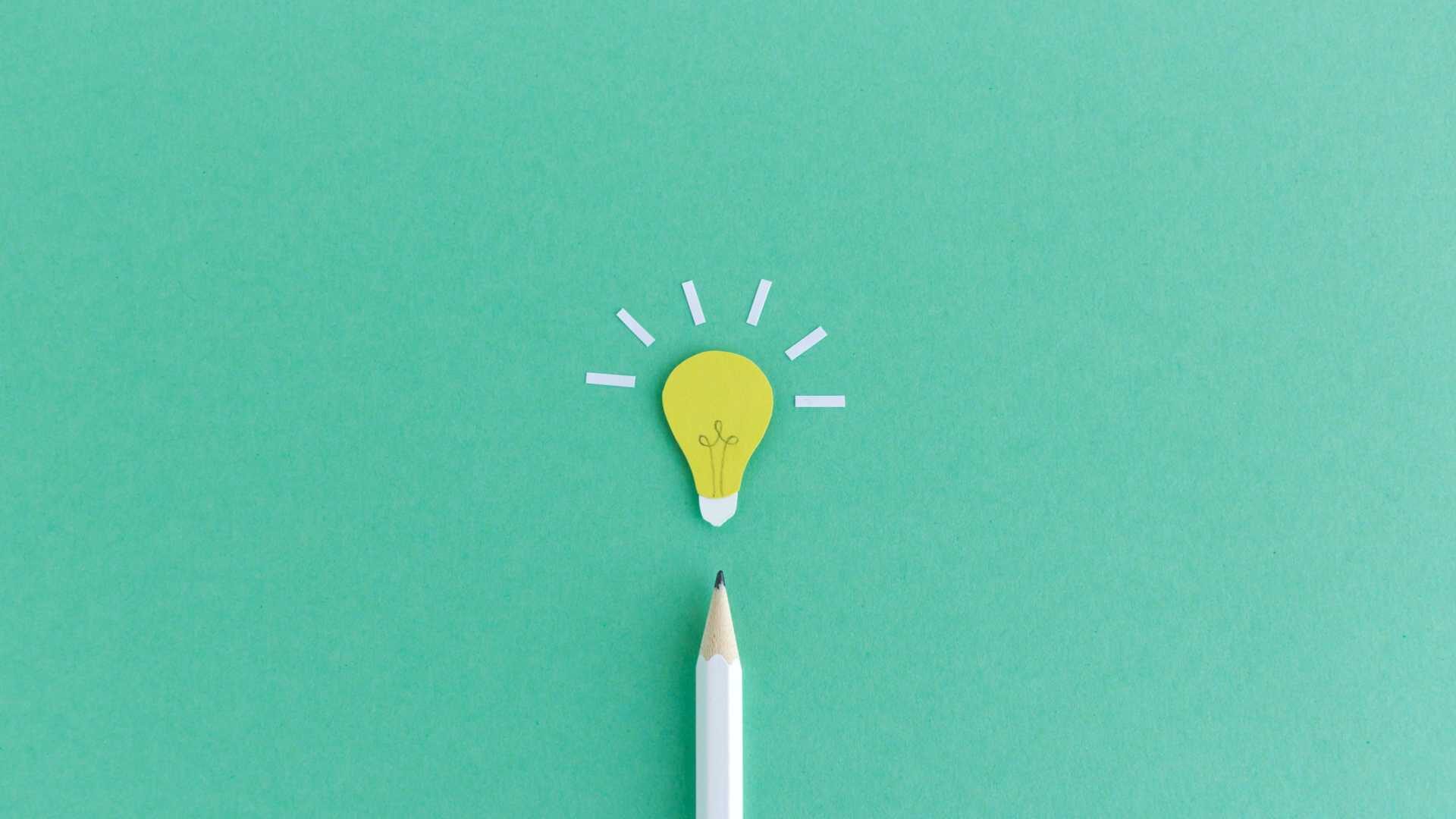 a white pencil points up from the bottom of the image at a small paper cutout of a lightbulb