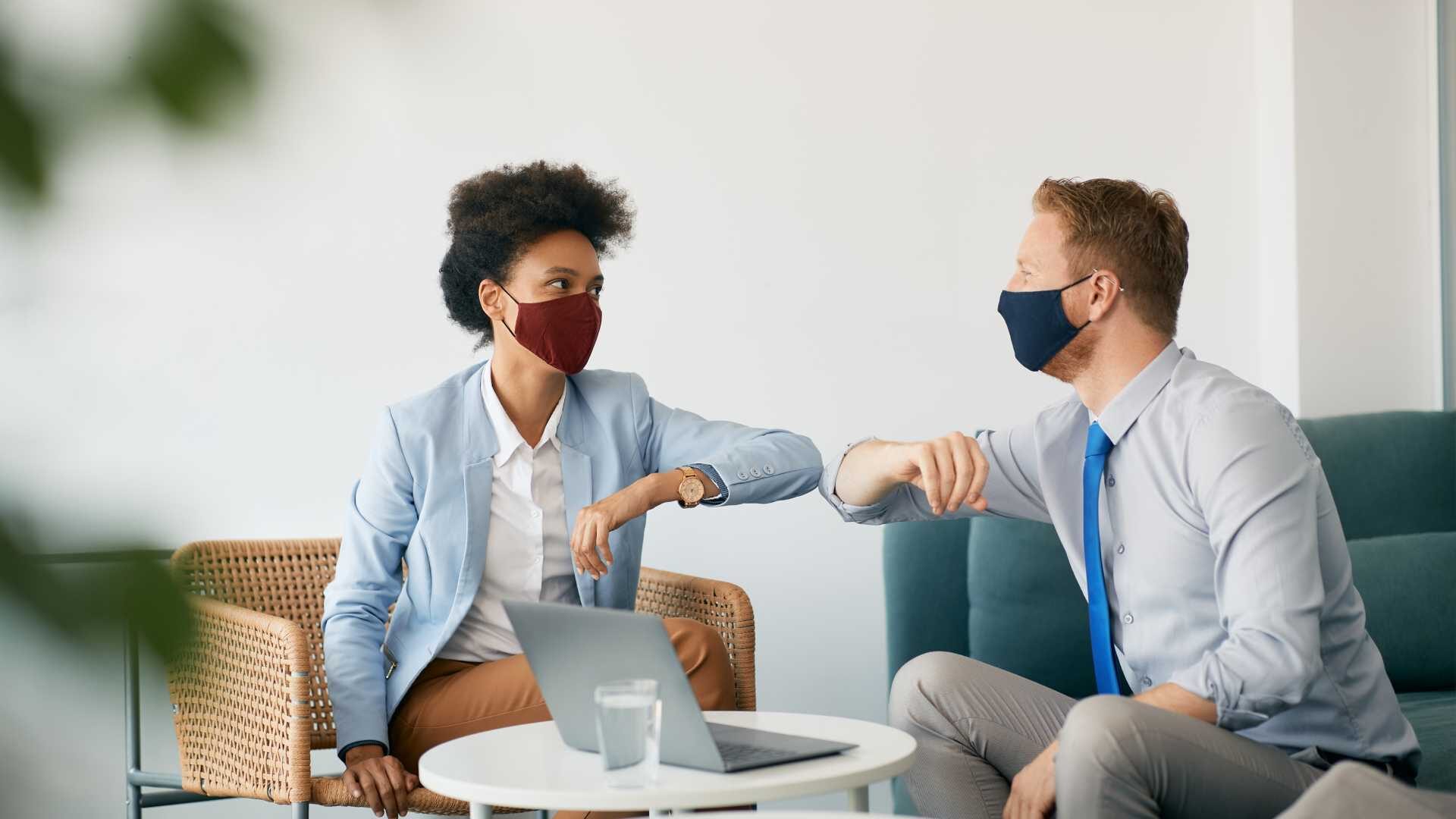 Two young professionals elbow bump while wearing masks in an office setting