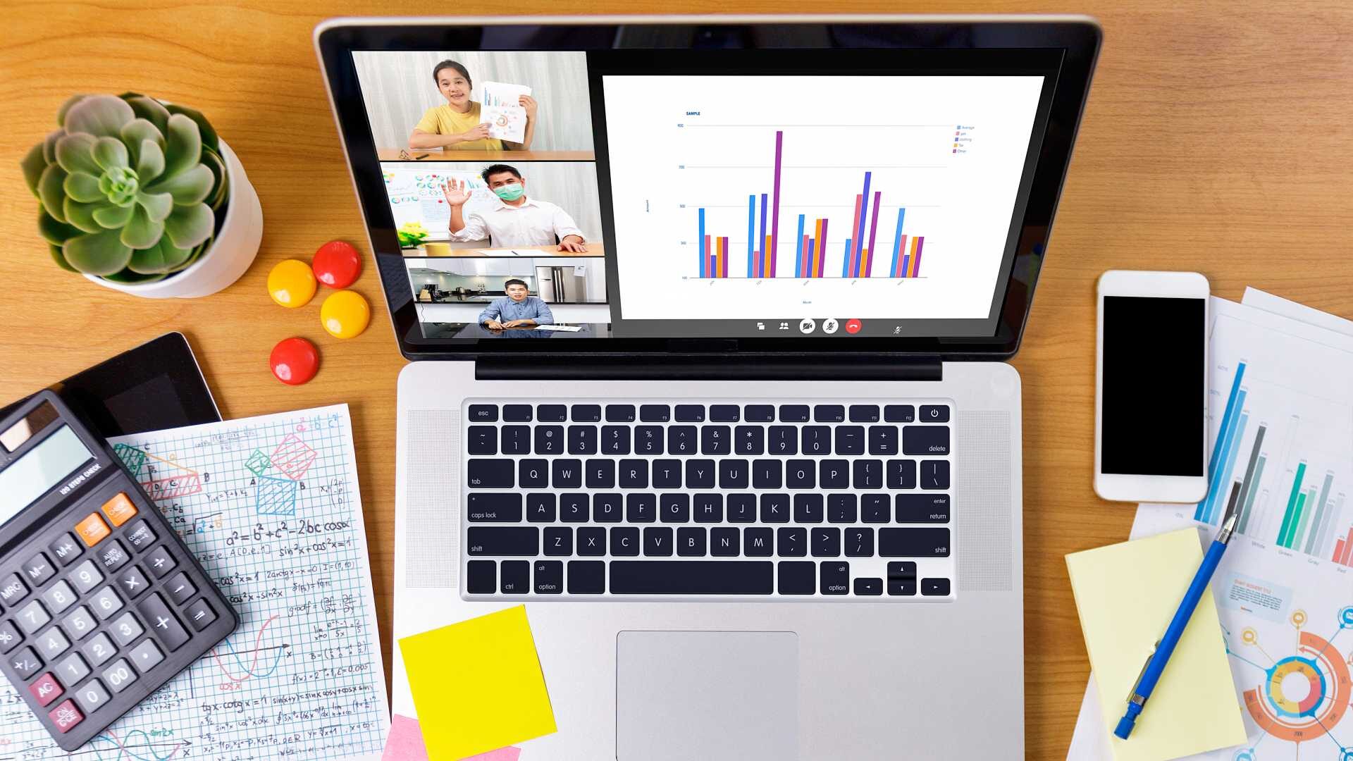 A desk viewed from above, with printouts, sticky notes and a smartphone spread around a laptop. The laptop screen shows coworkers discussing a slide of data and analytics.