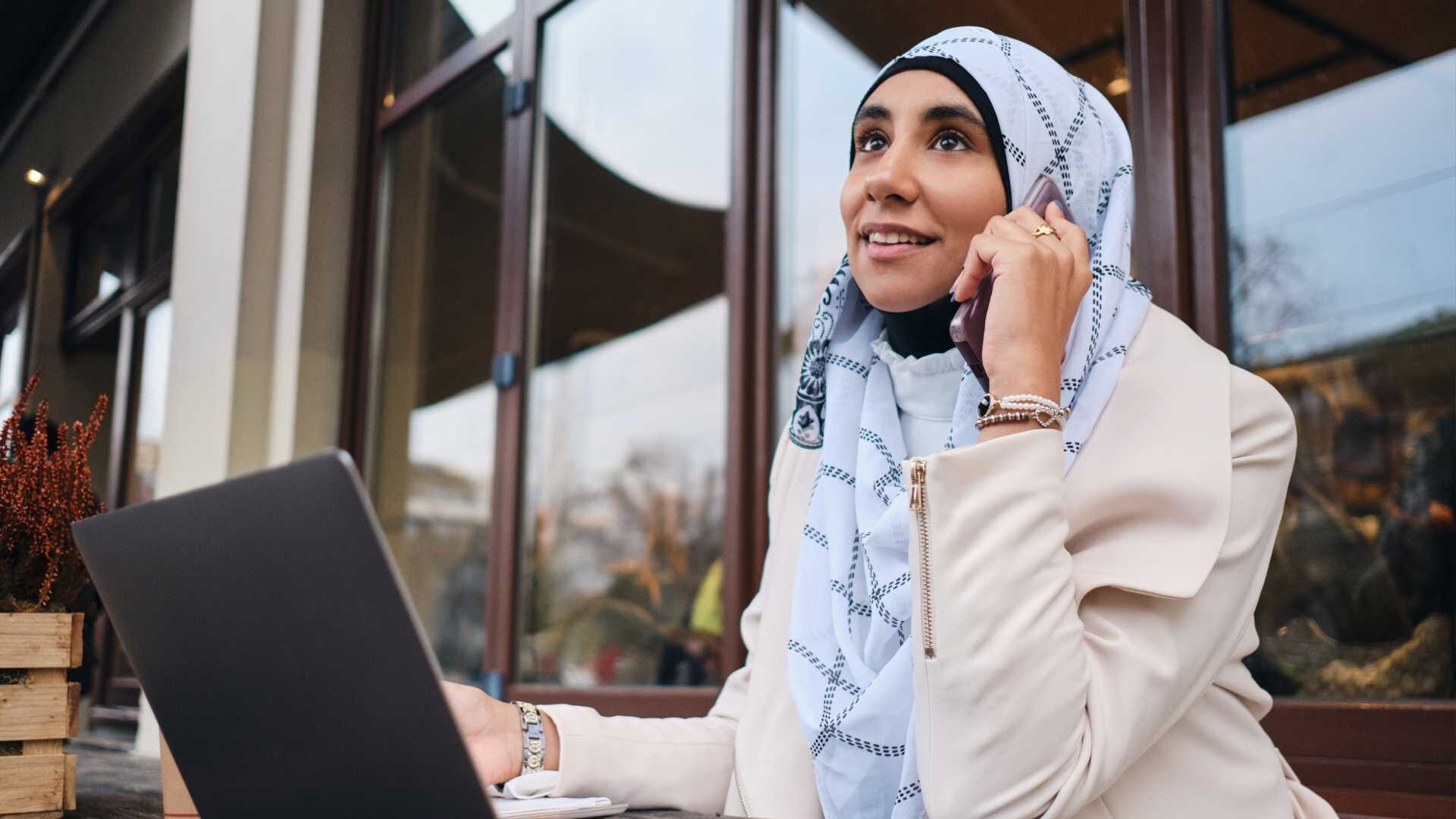 A young business woman in a headscarf works outdoors on her laptop.