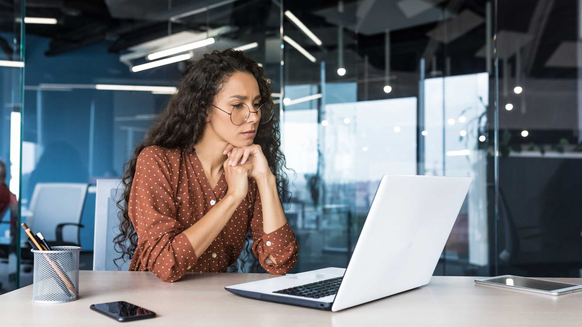 A business woman with glasses looks pensively at her laptop.