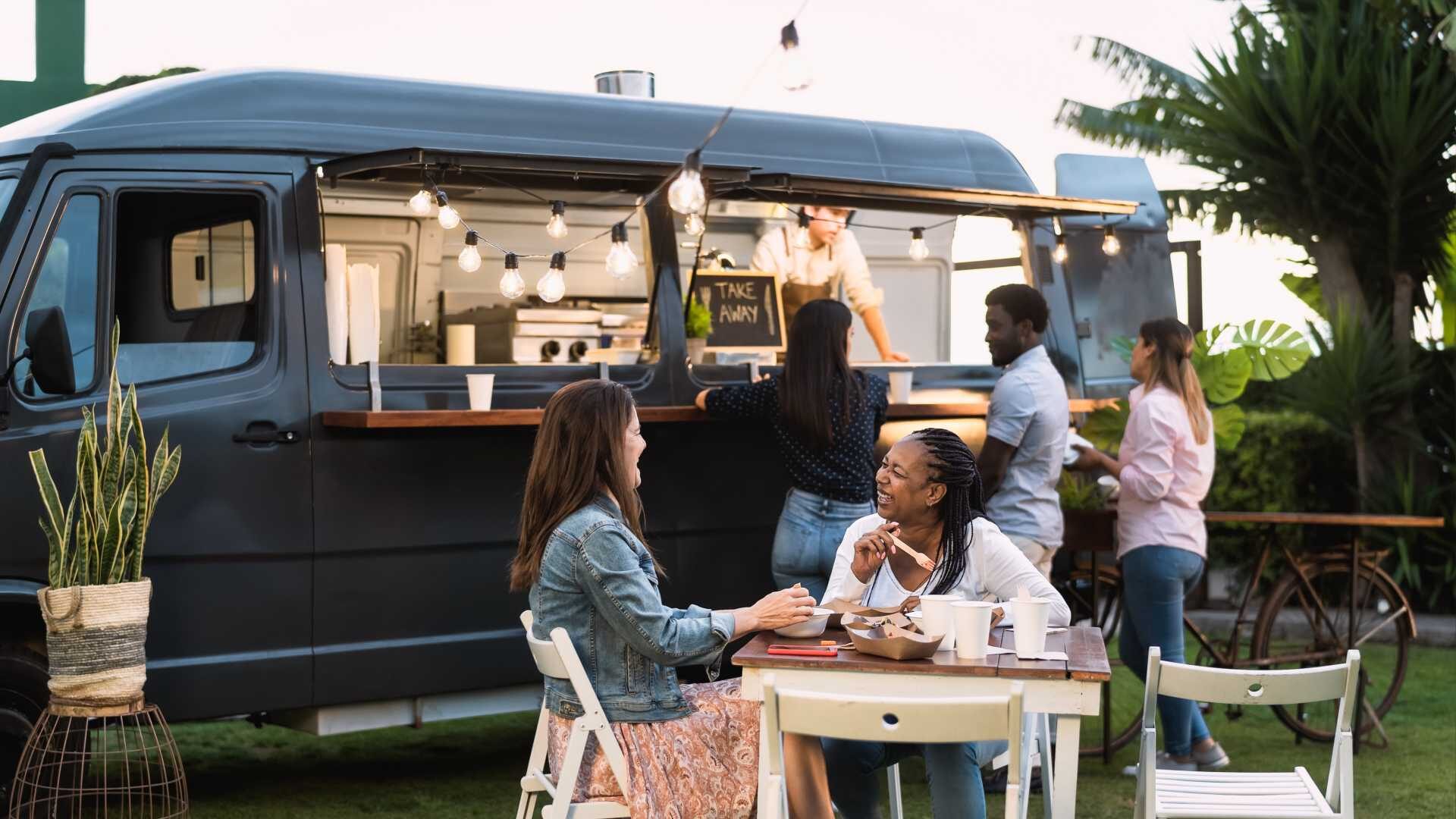 A diverse group of friends enjoys a meal in front of a food truck in the early evening.
