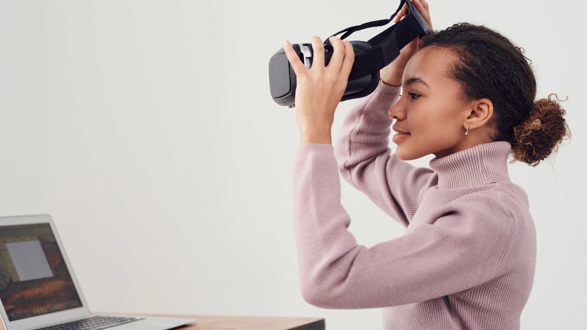 a woman puts on a virtual reality headset while seated at a desk in front of a laptop.