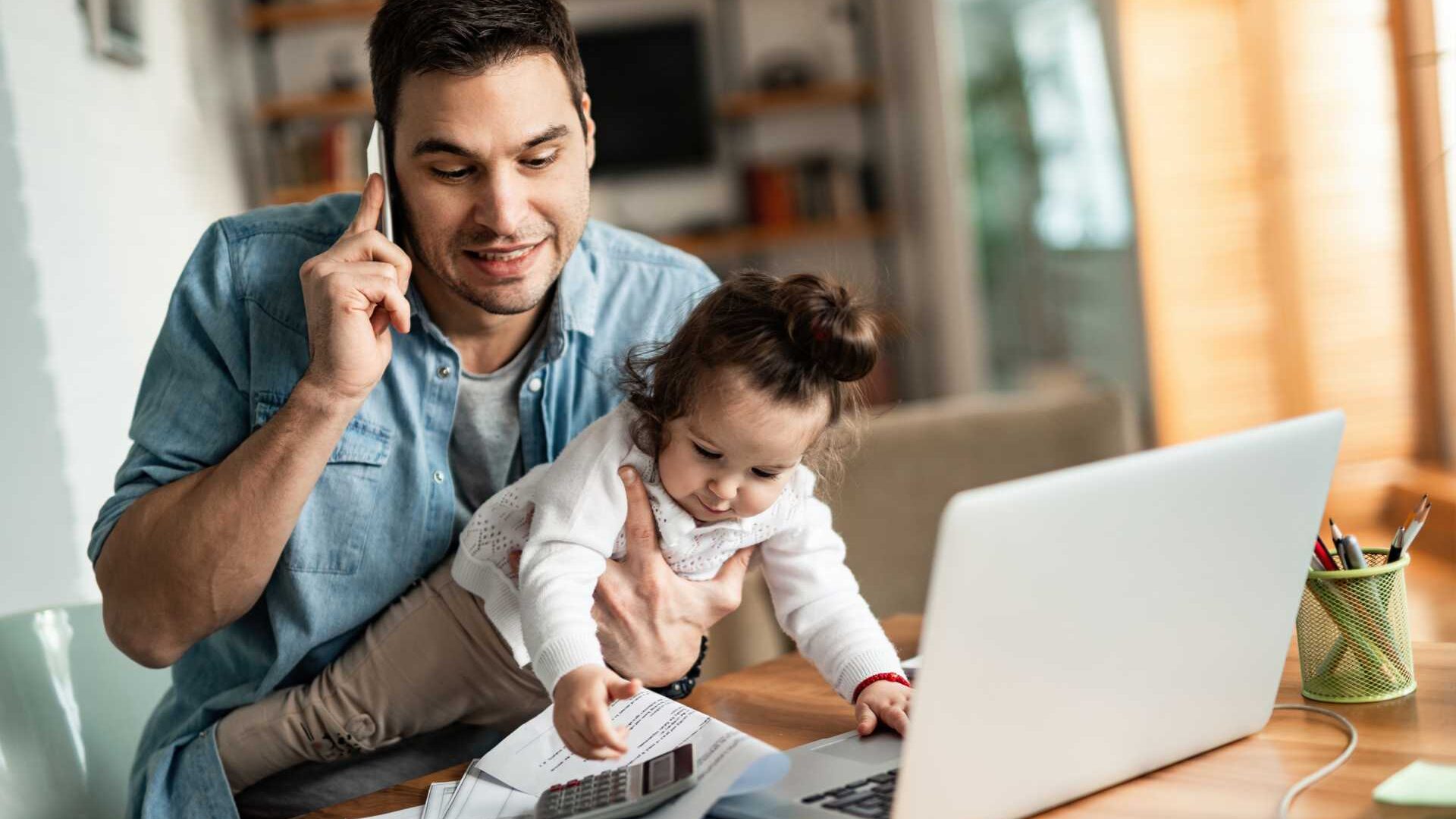 A working father sits at a desk him a home office with a toddler on his lap while talking on the phone.