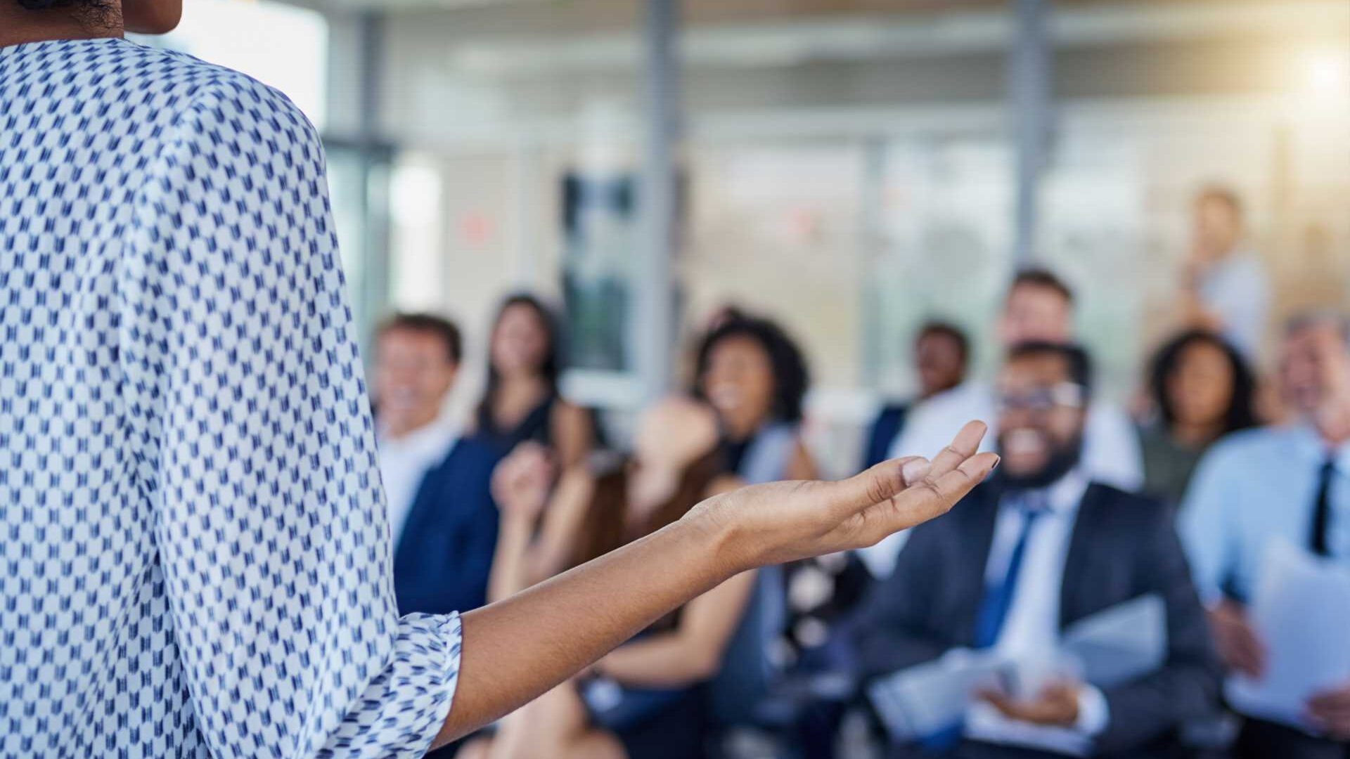A woman stands in front of an audience speaking in a professional setting