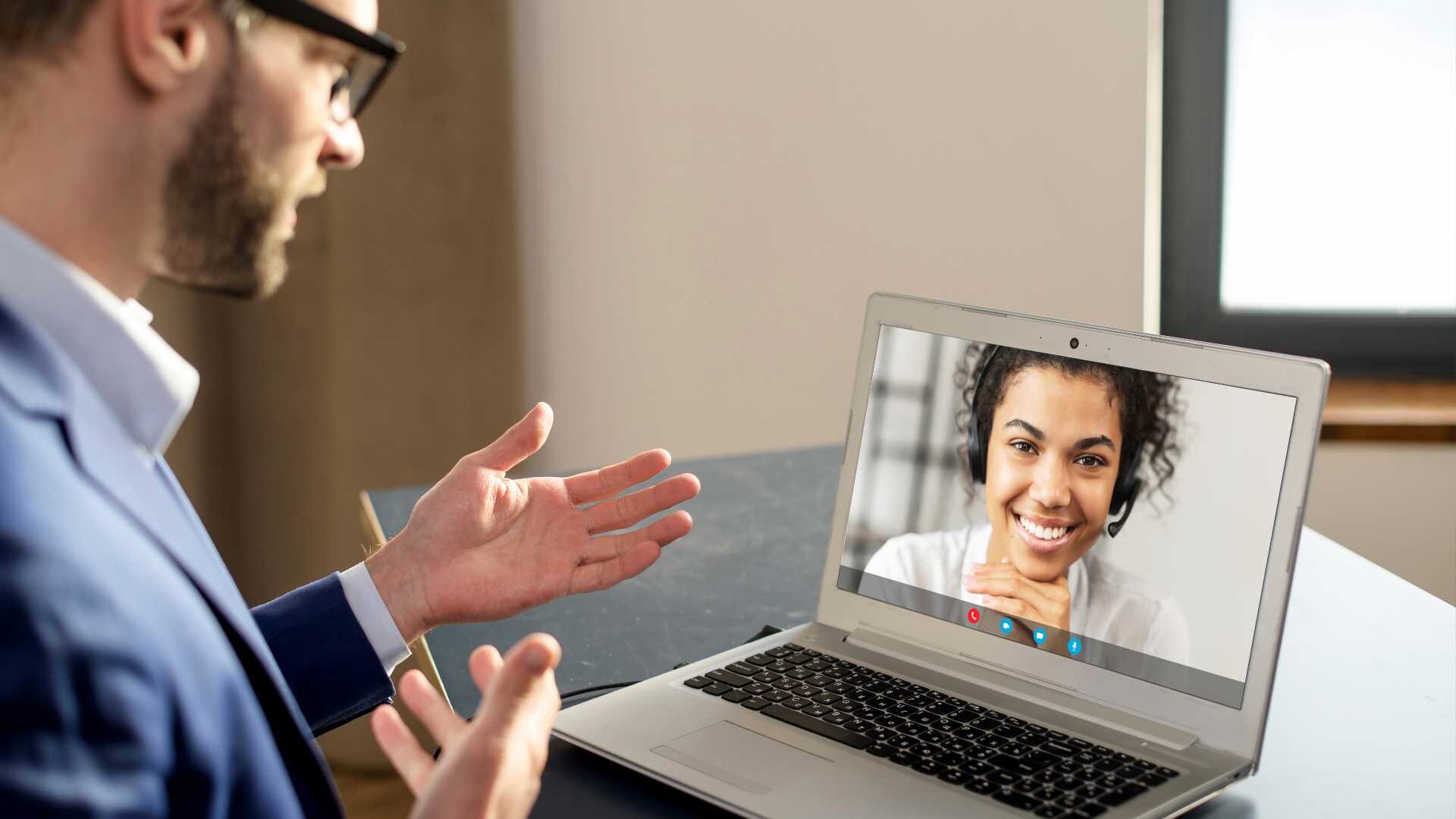 A man sits a desk and converses with a woman via video call in a business setting.