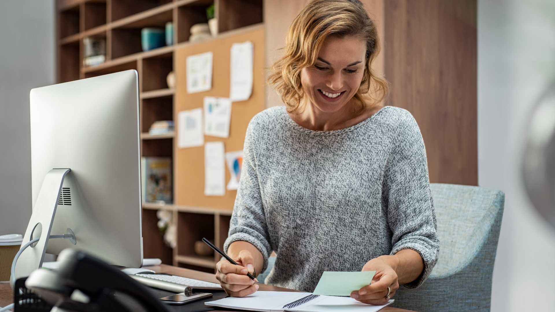 A smiling employee sits at a desk in front of a monitor while looking down at a notebook.