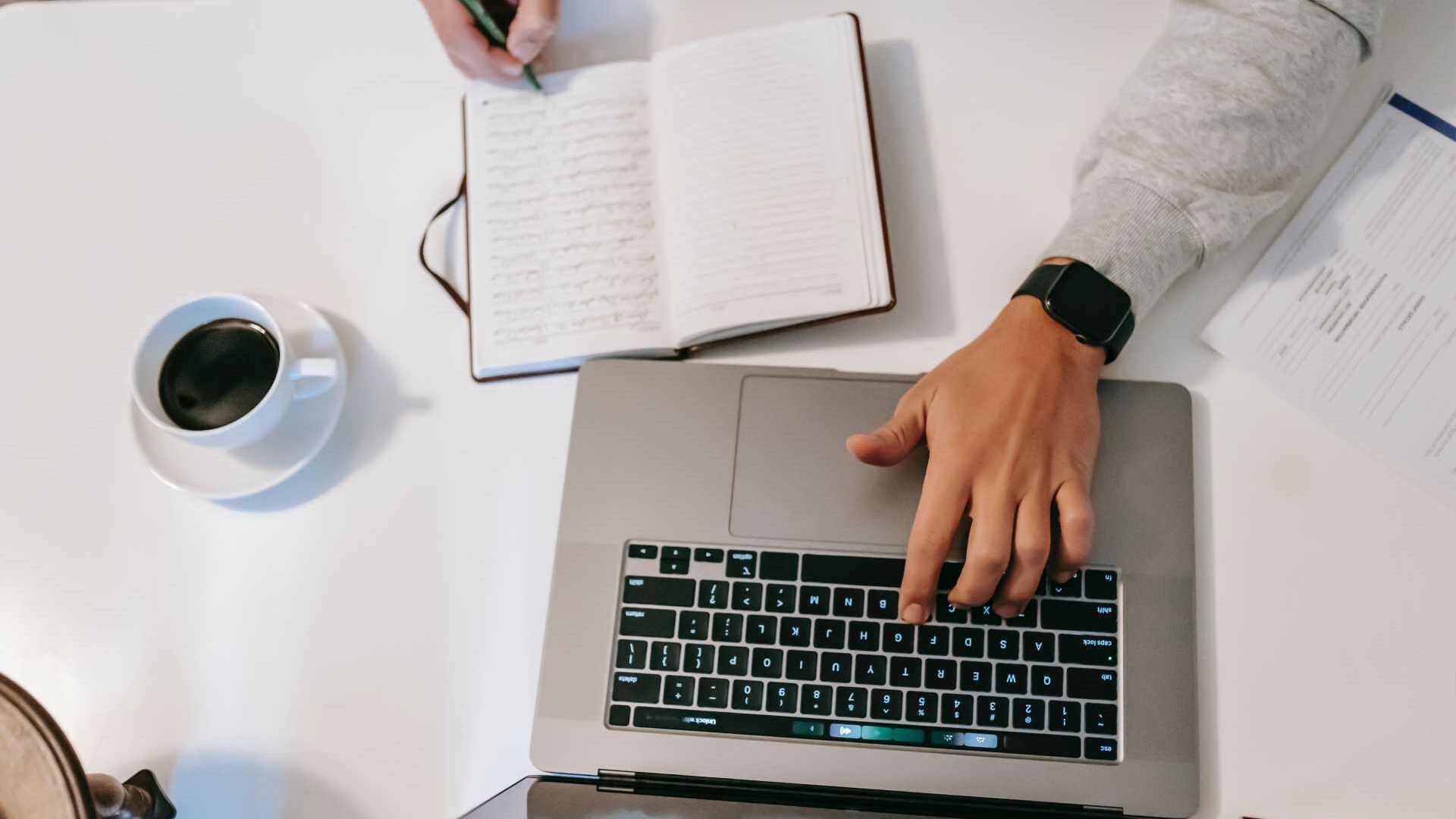 A hand rests on a laptop keyboard next to an open notebook with a cup of coffee placed on a white table.