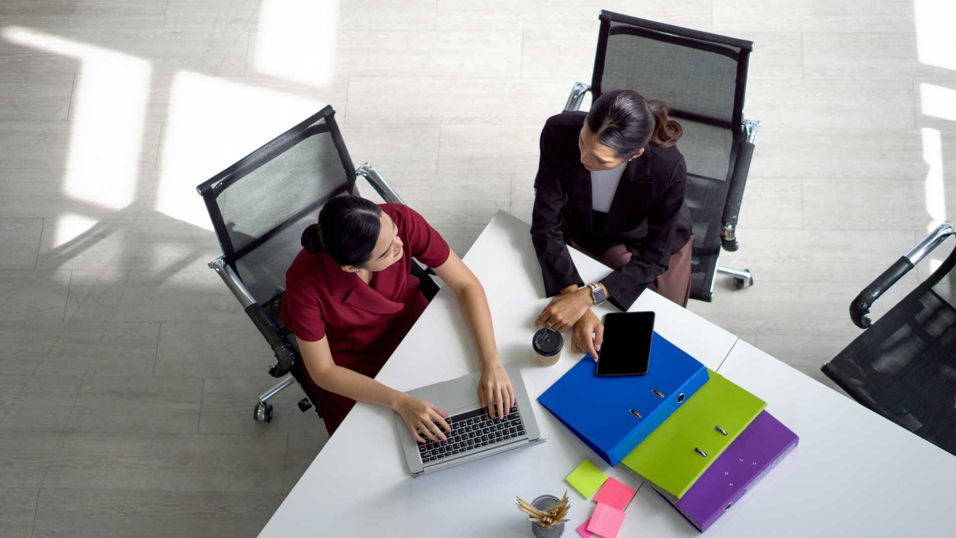 Two women sit at a conference table in an office setting in front of a laptop.