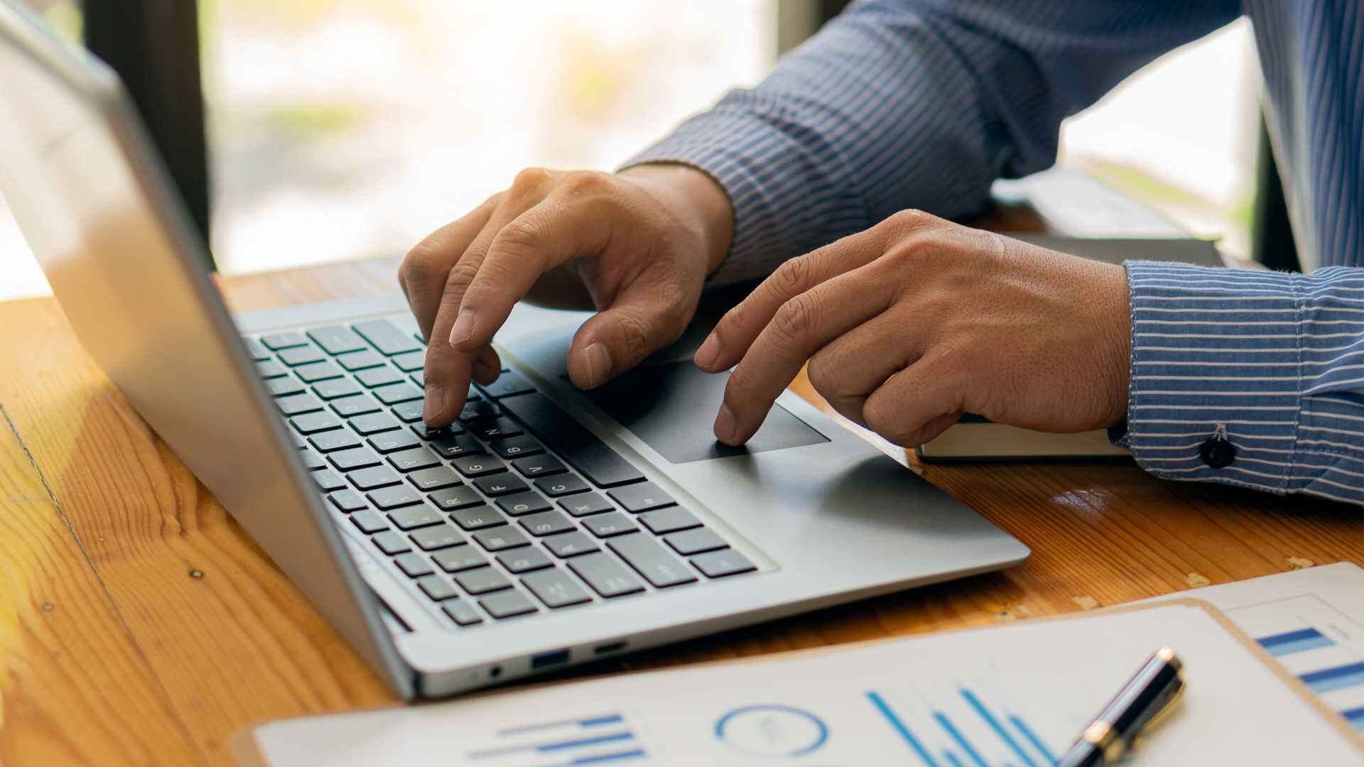 A person types on a laptop sitting on a desk beside a paper with charts and graphs.