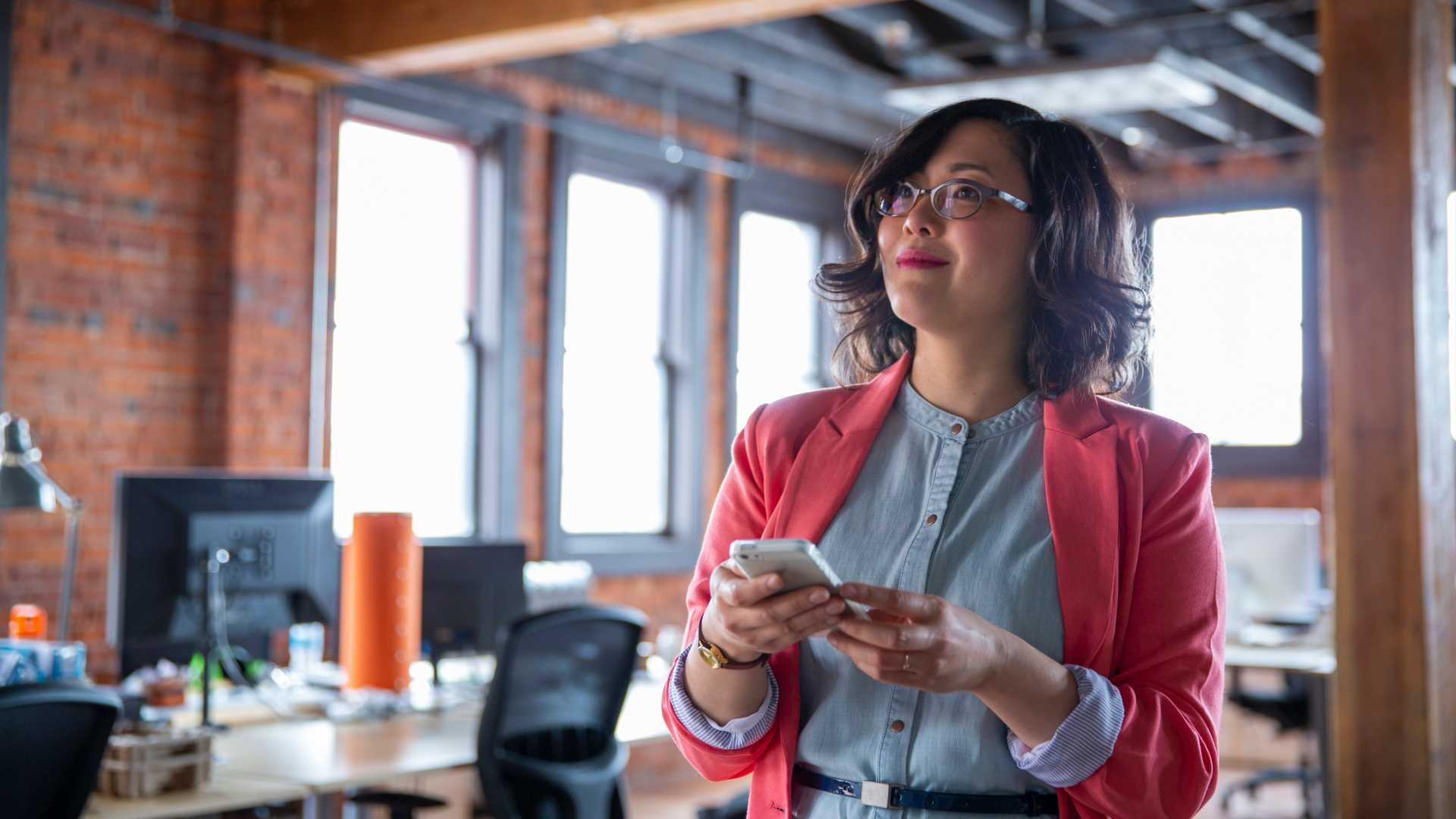A woman in a professional setting holds a mobile phone while looking into the distance.