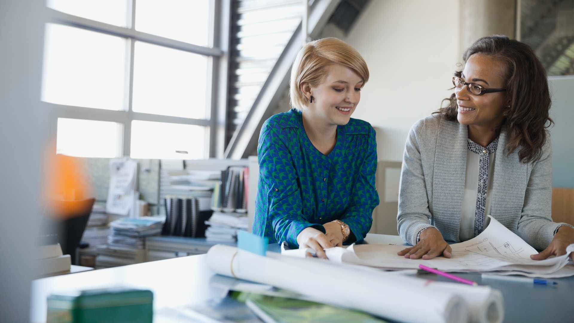Two women work together in a professional setting by a sunny window.