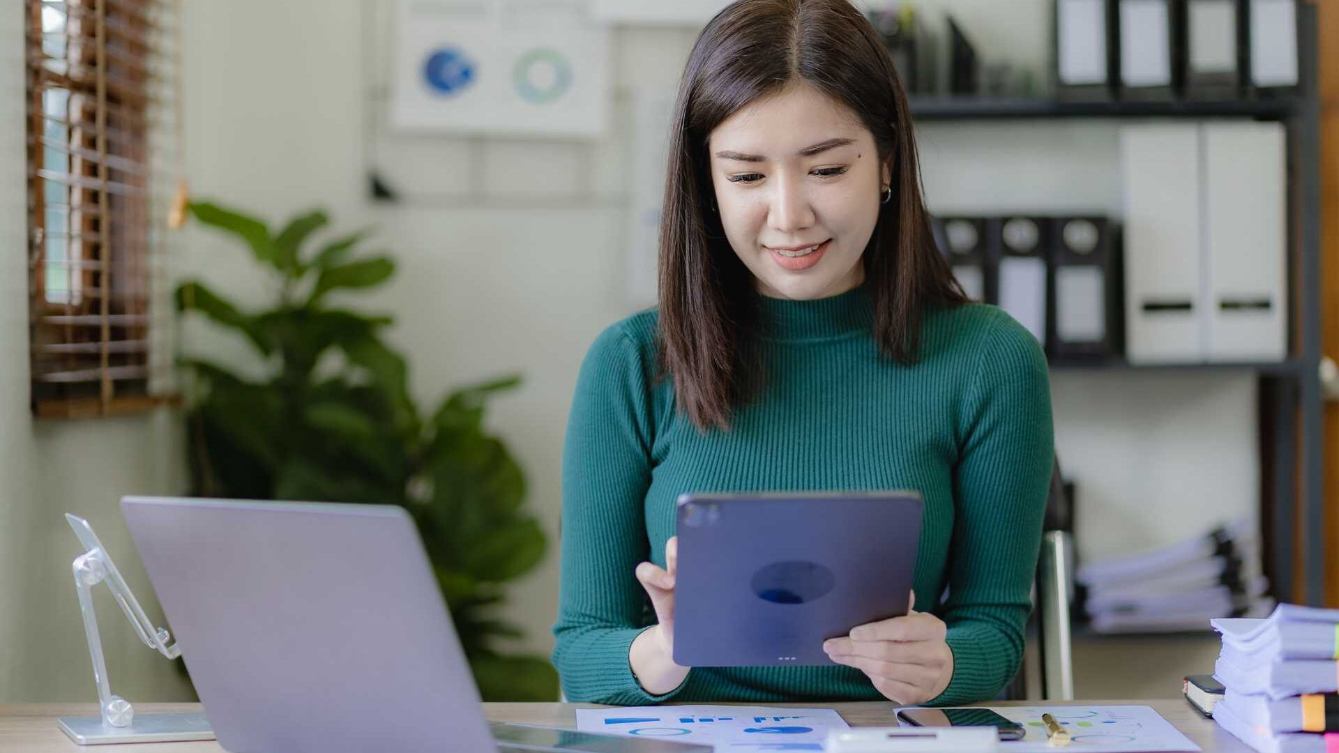 A woman sits at a desk in front of a laptop while using a tablet.