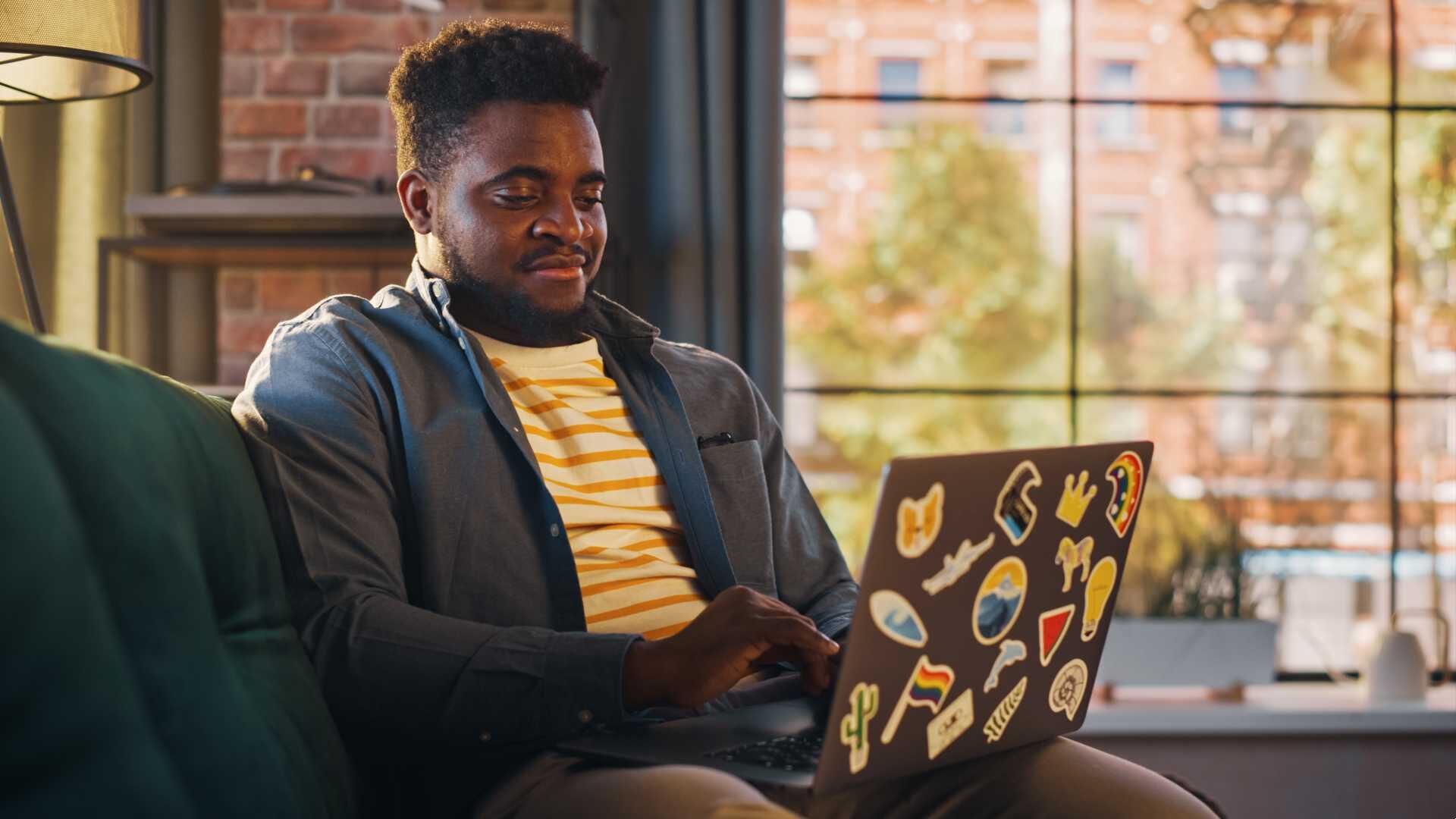 A young Black professional works from home on his laptop, which is covered in stickers.