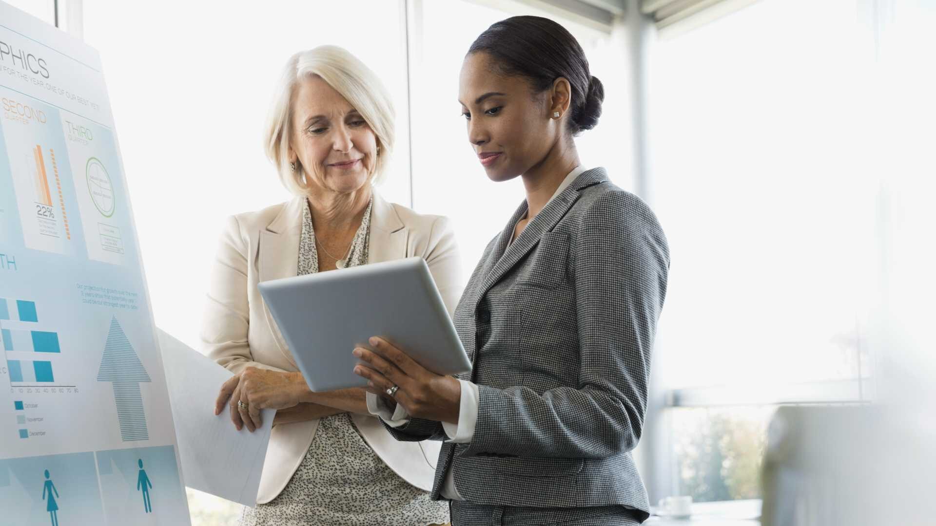 Two business people stand in front of a sales chart while looking at a tablet.
