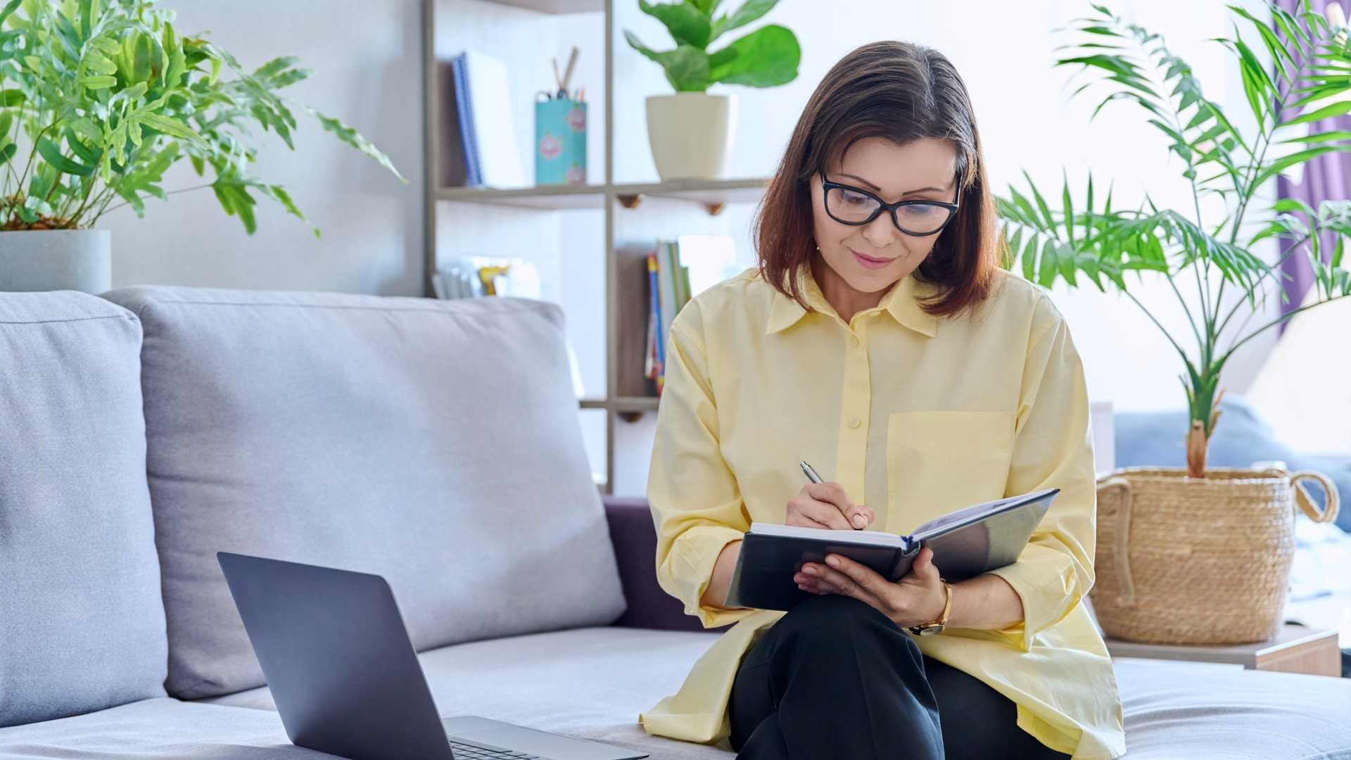 A woman writes in a notebook while sitting on a couch in front of a laptop