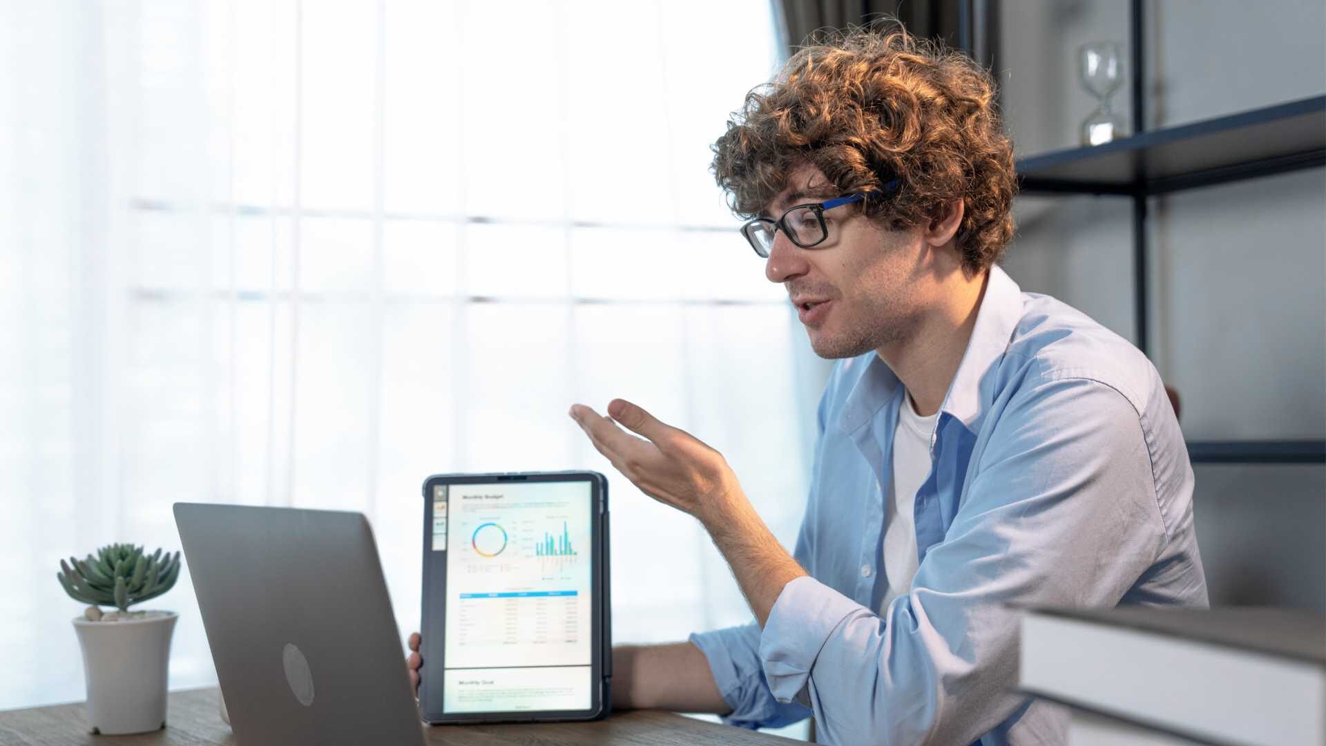 A young businessman is explaining cybersecurity concepts to a coworker in a video chat. He holds up a tablet, gesturing to various graphs.