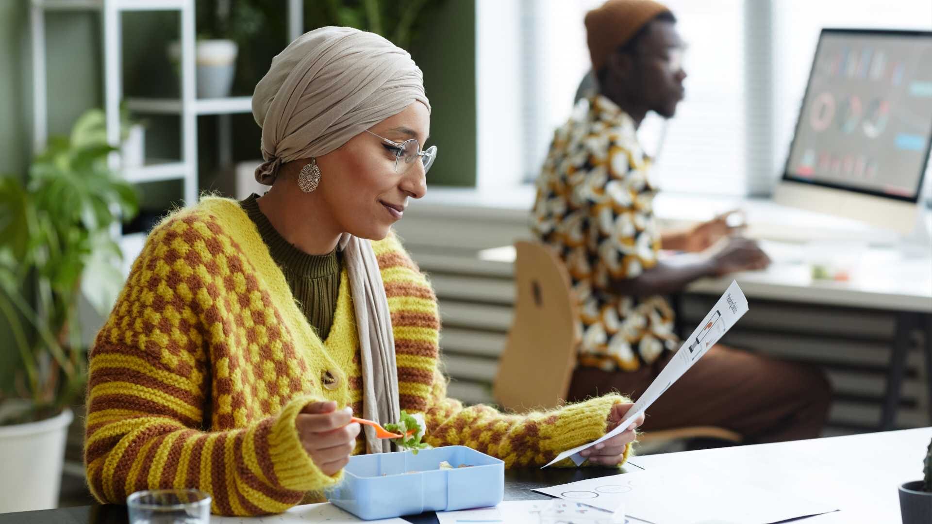 Woman eating a light salad at her desk while reviewing a document. Her colleague is in the background working on the computer.