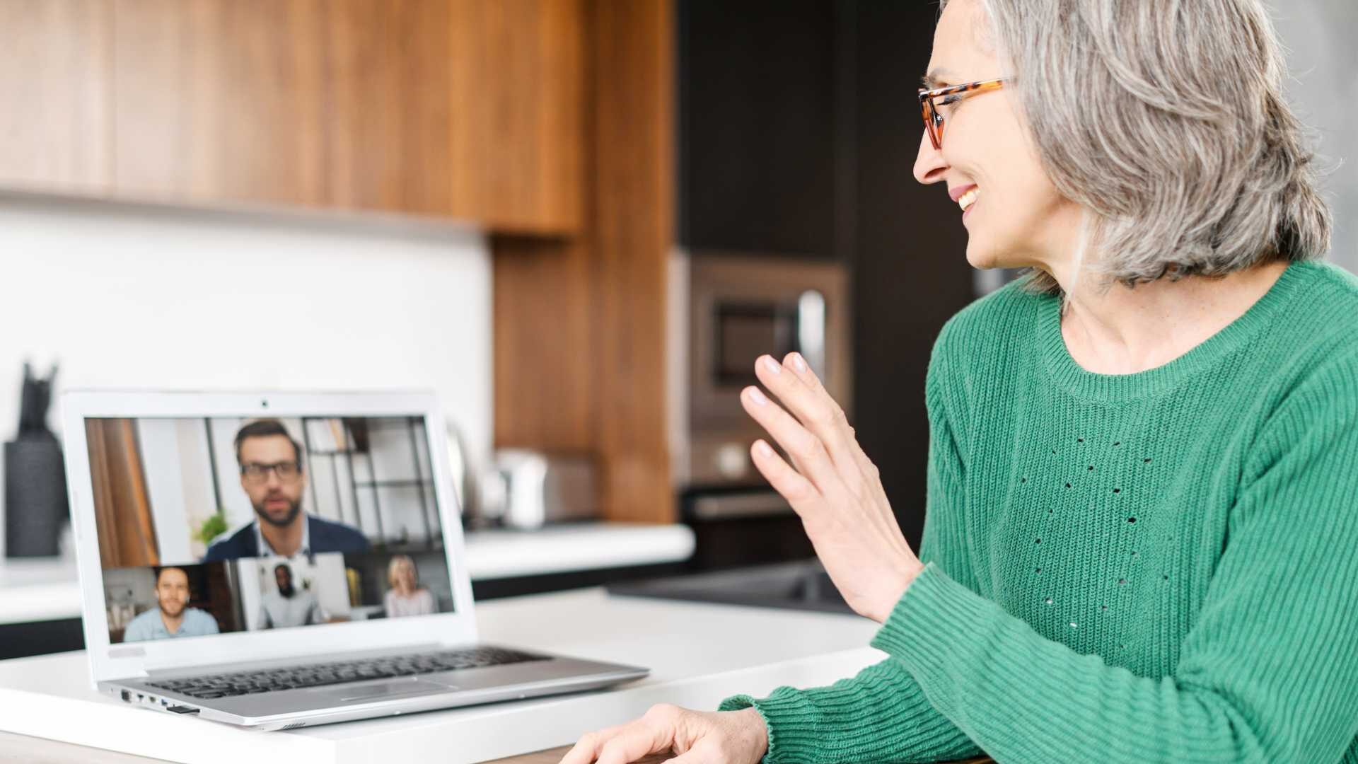 A woman is sitting at a laptop in a virtual meeting, waving to her coworkers