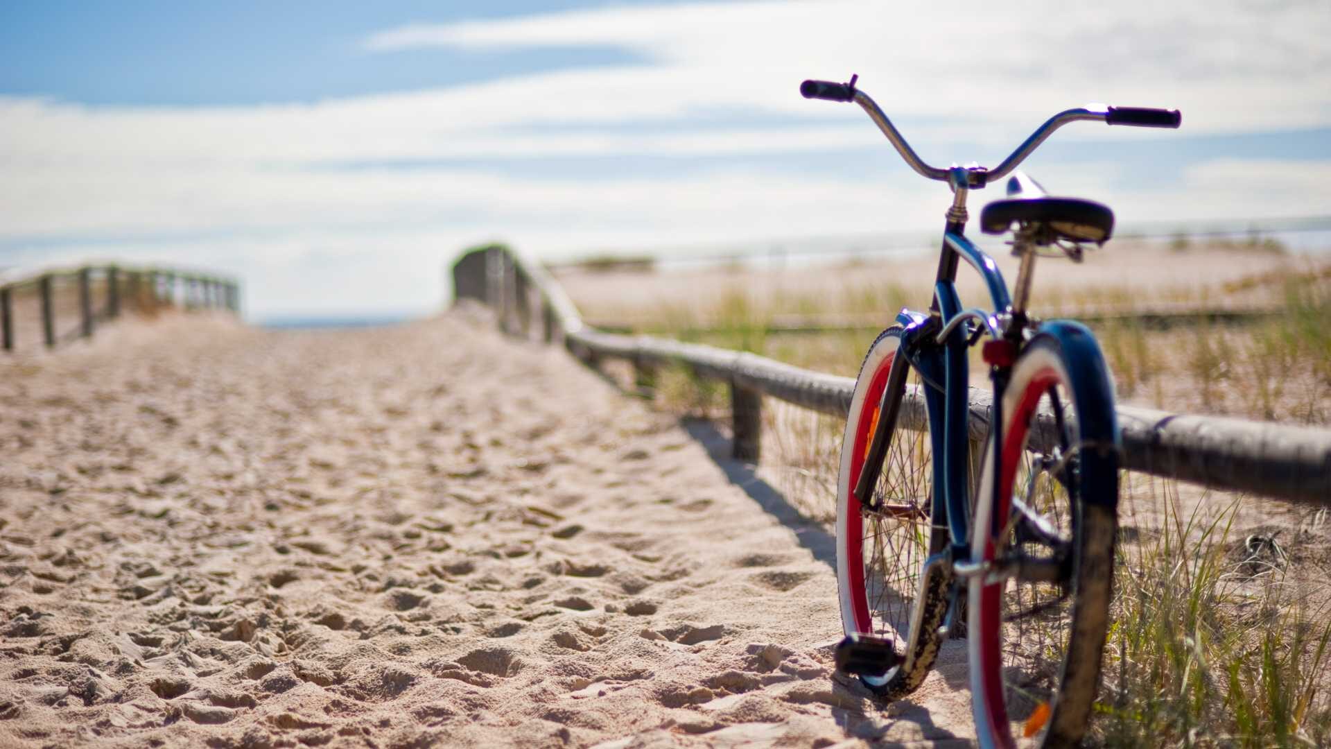 A bicycle leans on a wooden railing. It is sitting on a sandy path that leads to the beach on a sunny day.