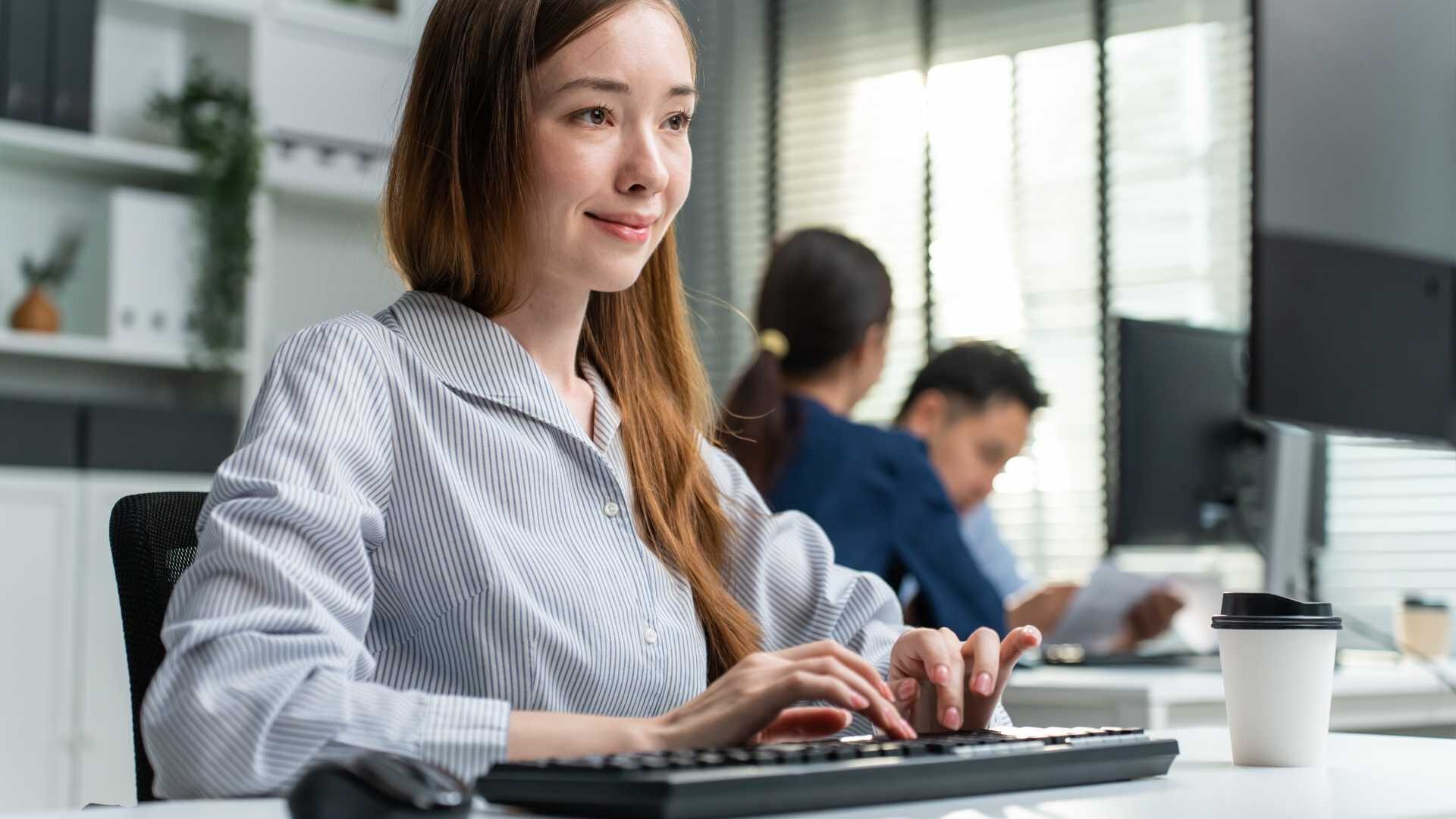 Young woman sitting at a desk in a striped blue button up shirt typing on a keyboard with a small coffee cup at her desk.