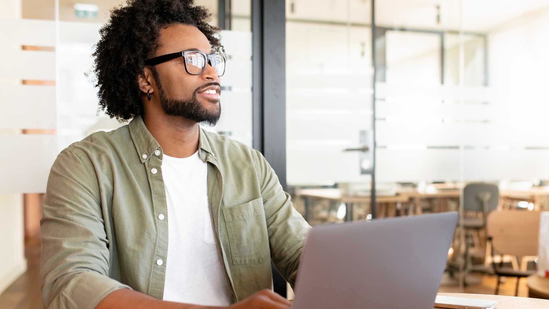 A young professional looks thoughtfully up from his laptop.