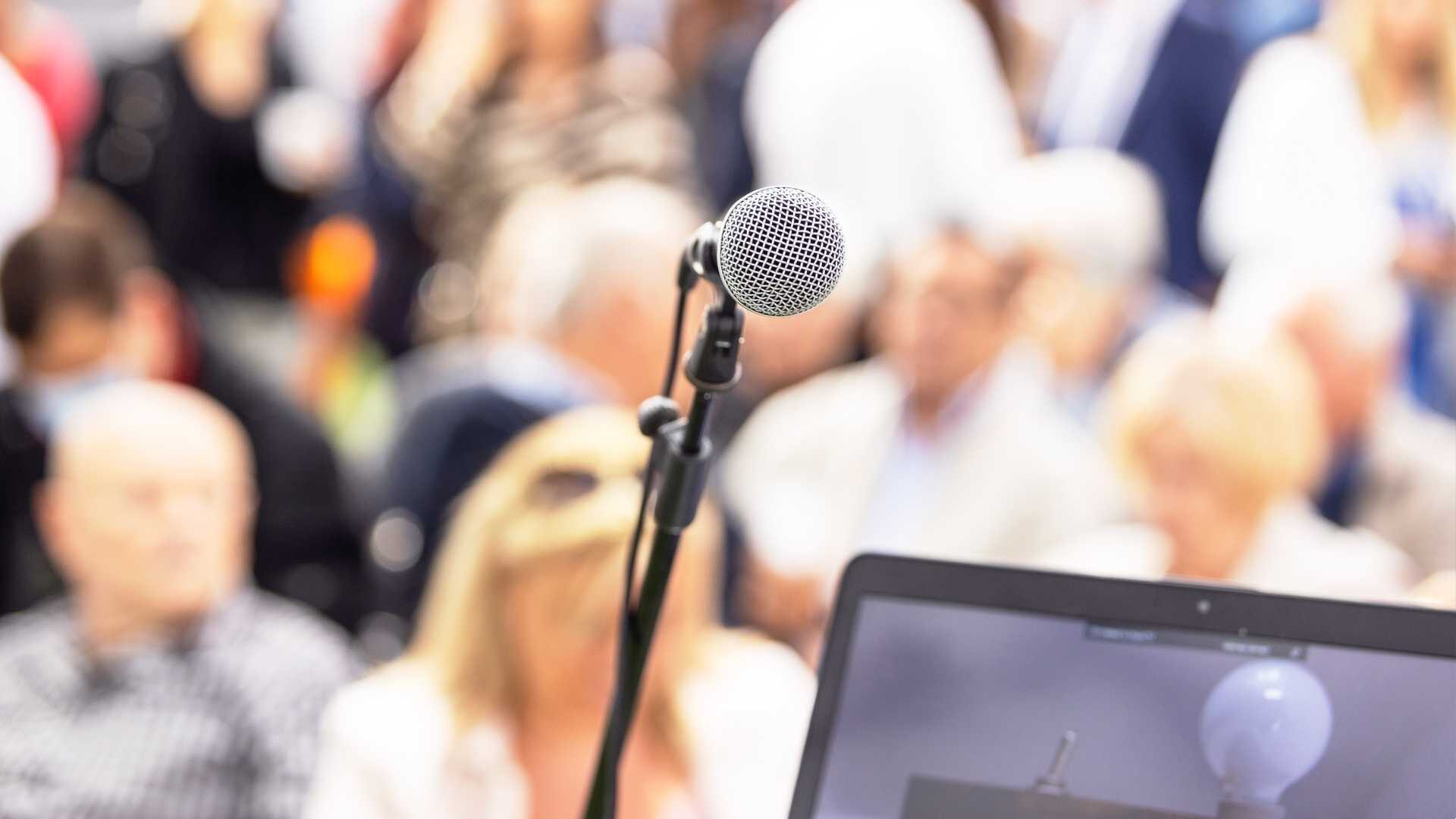 Microphone on stage in front of an audience of people, laptop in the corner of the camera shot.