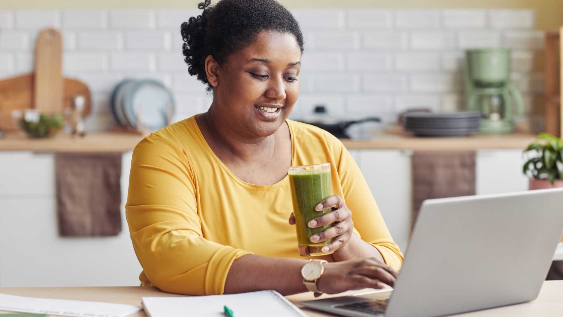 A woman drinking a green smoothie works from home in front of a laptop