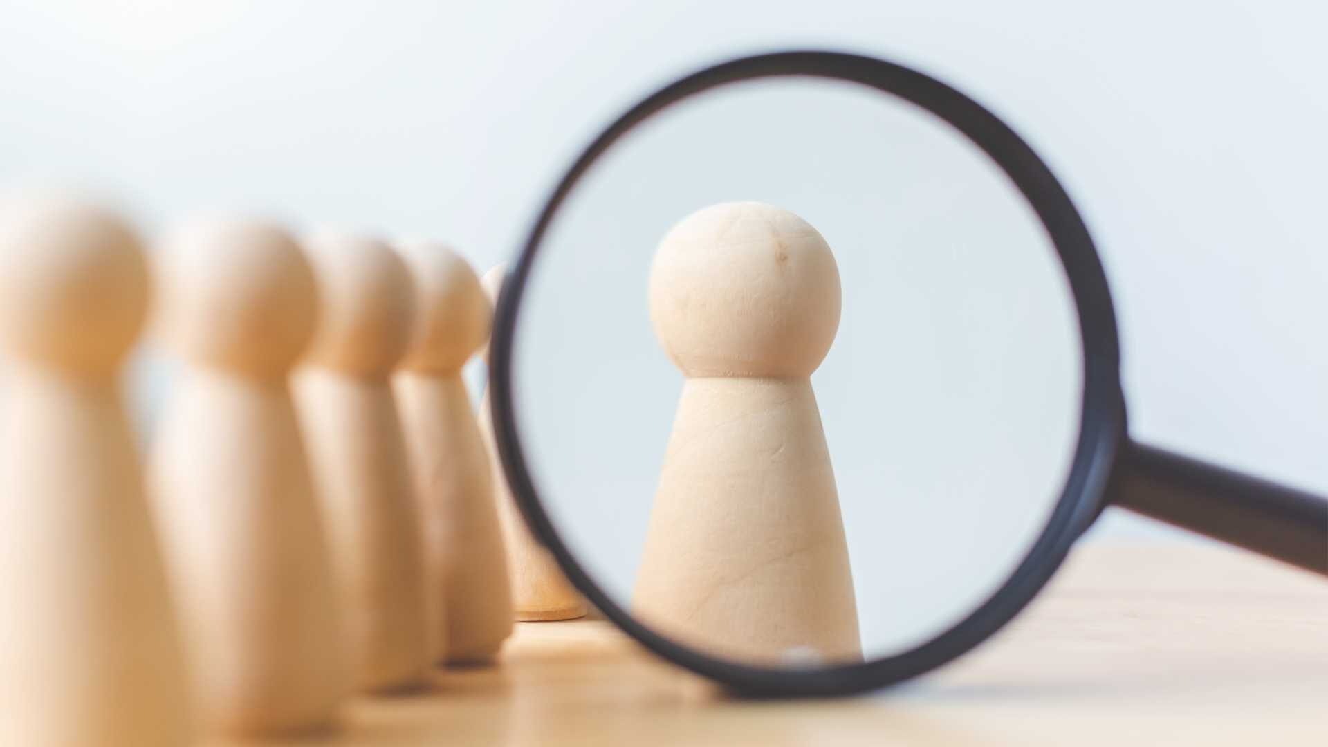 Wood pawns in a row with a magnifying glass observing one of the pawns.