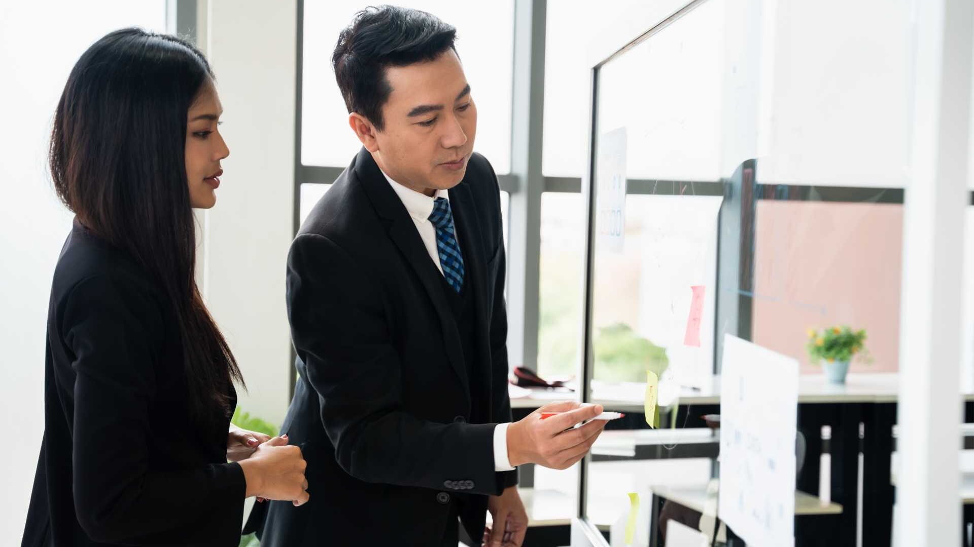 To employees in formalwear collaborate in front of a whiteboard in an office space