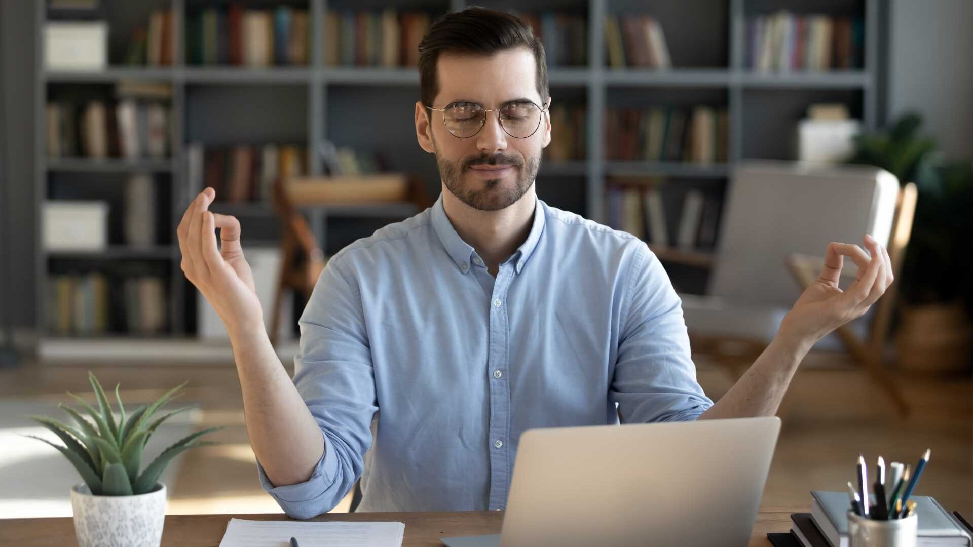 A man sits at a desk in front of a laptop while meditating