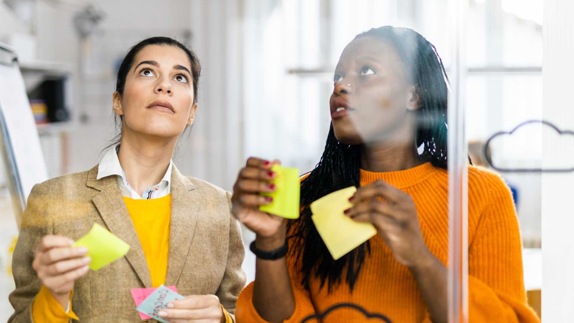 Two workers collaborating at a glass board with sticky post it notes.