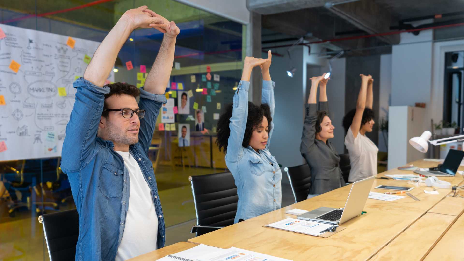 Colleagues sitting at a desk, all of them stretching their arms above their heads. Their work is in front of them on the table.