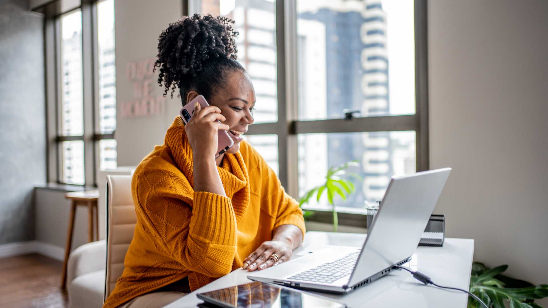 A businesswoman talks on her cell phone happily while looking at her laptop screen. She's probably looking at personalized sales training content.