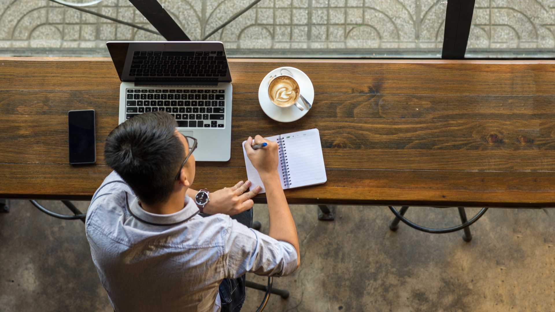 A top-down view of a young professional sitting at a cafe bar and writing notes in a notebook with his laptop open in front of him and a latte next to it.