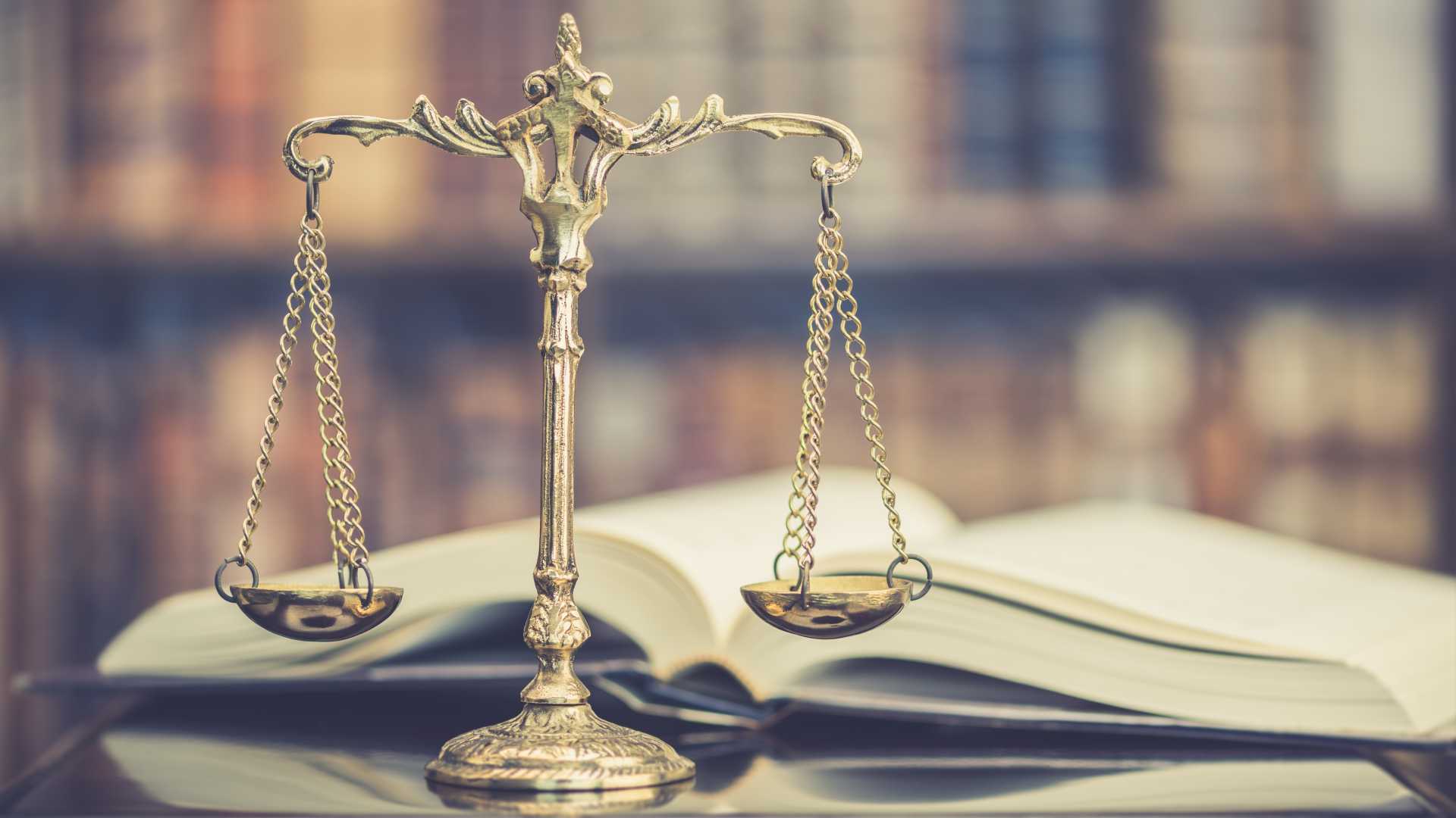 Justice scales on a formal professional judicial desk with an open book and bookshelf blurred in background.