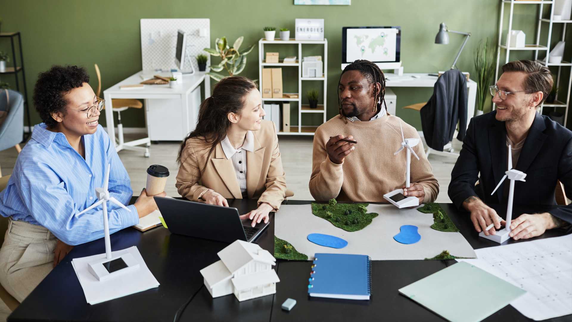 A group of coworkers sit around a table talking and smiling with model wind turbines in hand