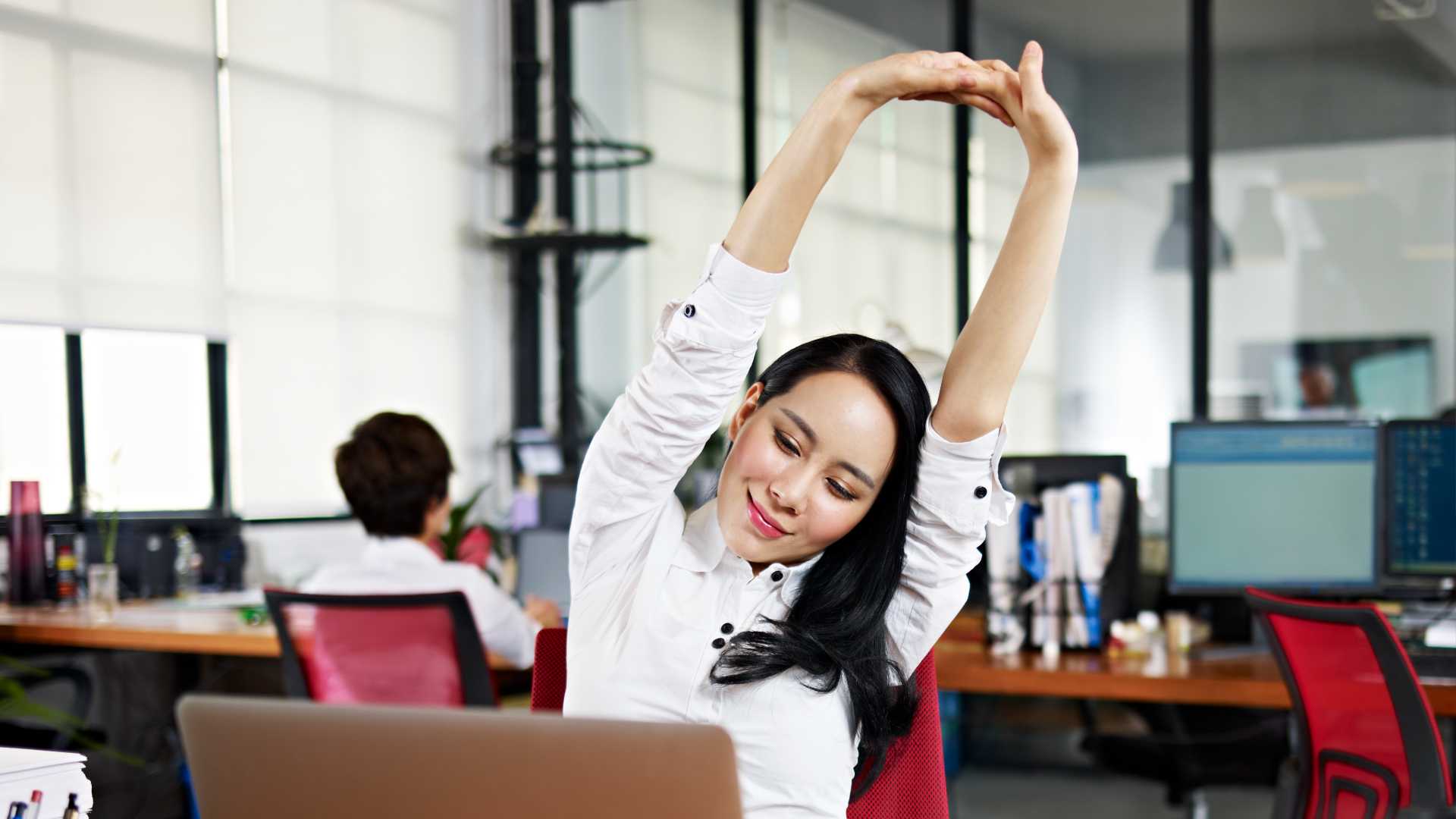 Colleague sitting at their desk stretching arms into the air with a small, relaxed smile.