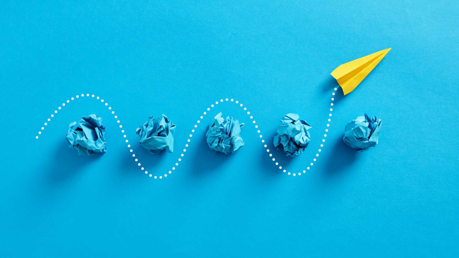 Blue background with crumpled blue CGI paper and a CGI yellow paper airplane leaving white tracks as it weaves through the paper balls.