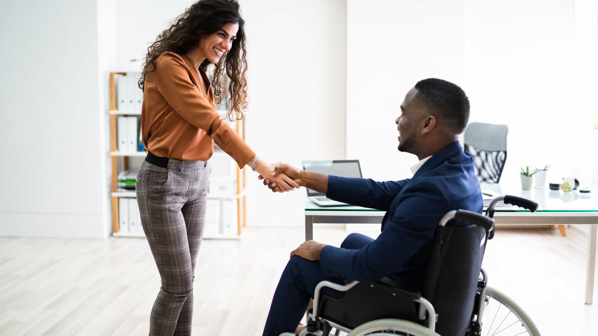 A man seated in a wheelchair shakes hands with a woman who is standing up in a professional setting