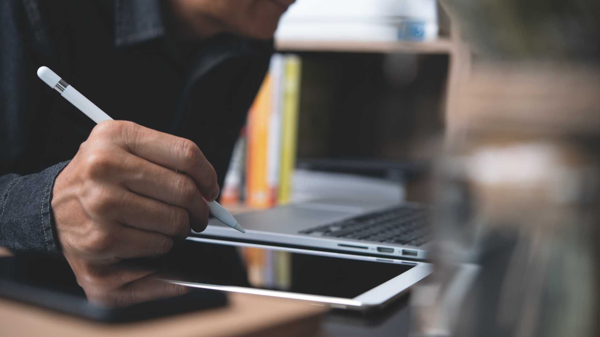A man works in front of a laptop while holding a digital pen and writing on a tablet