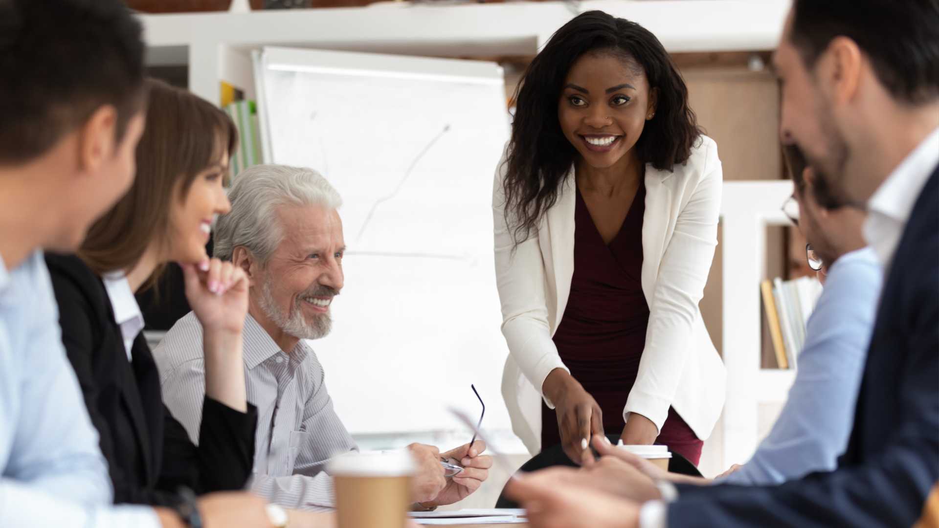 Co-workers are gathered in an office around a table. One is standing and smiling at everyone