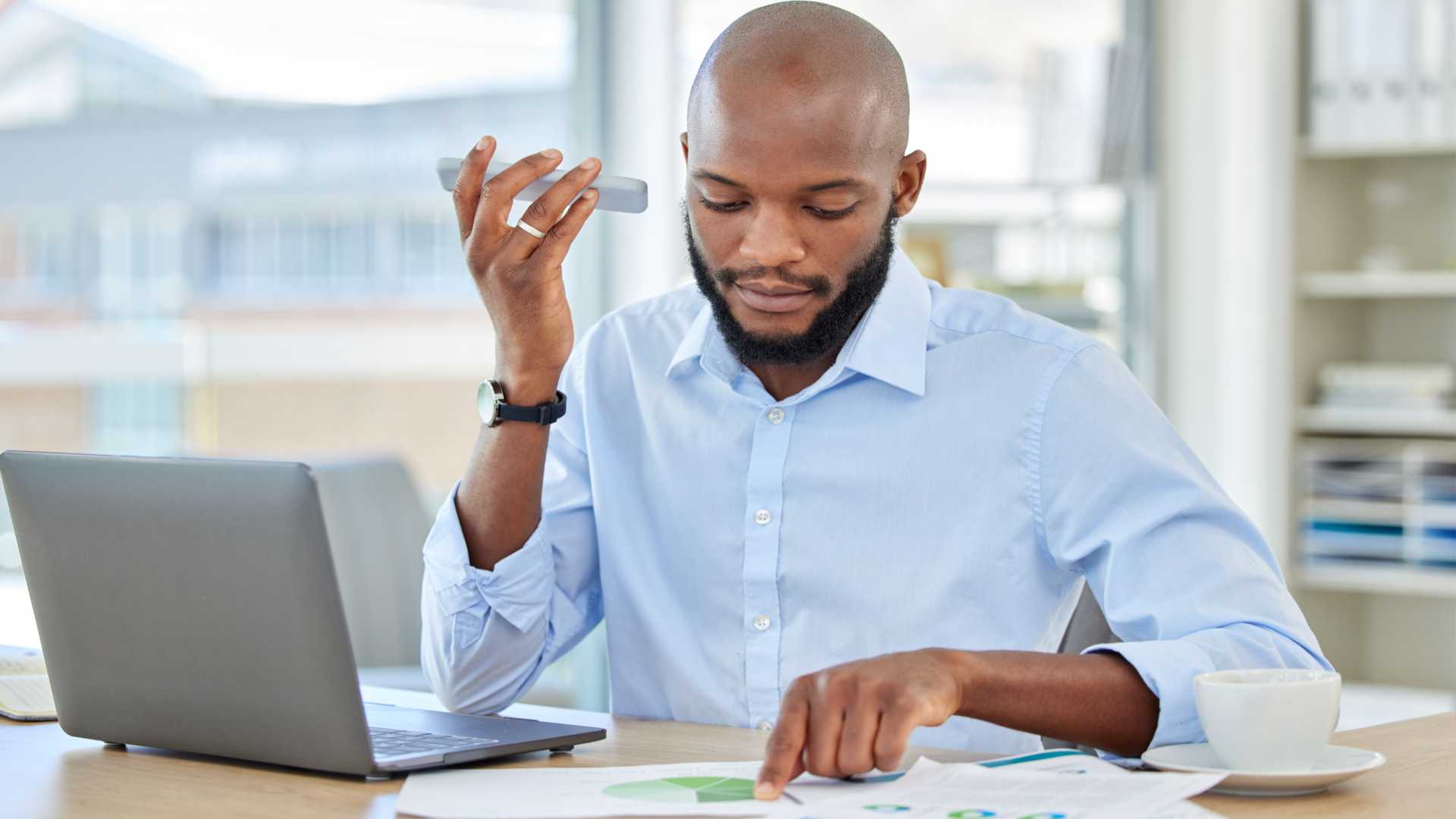 A man is sitting in front of computer while holding a cell phone and looking at charts laid out on his desk